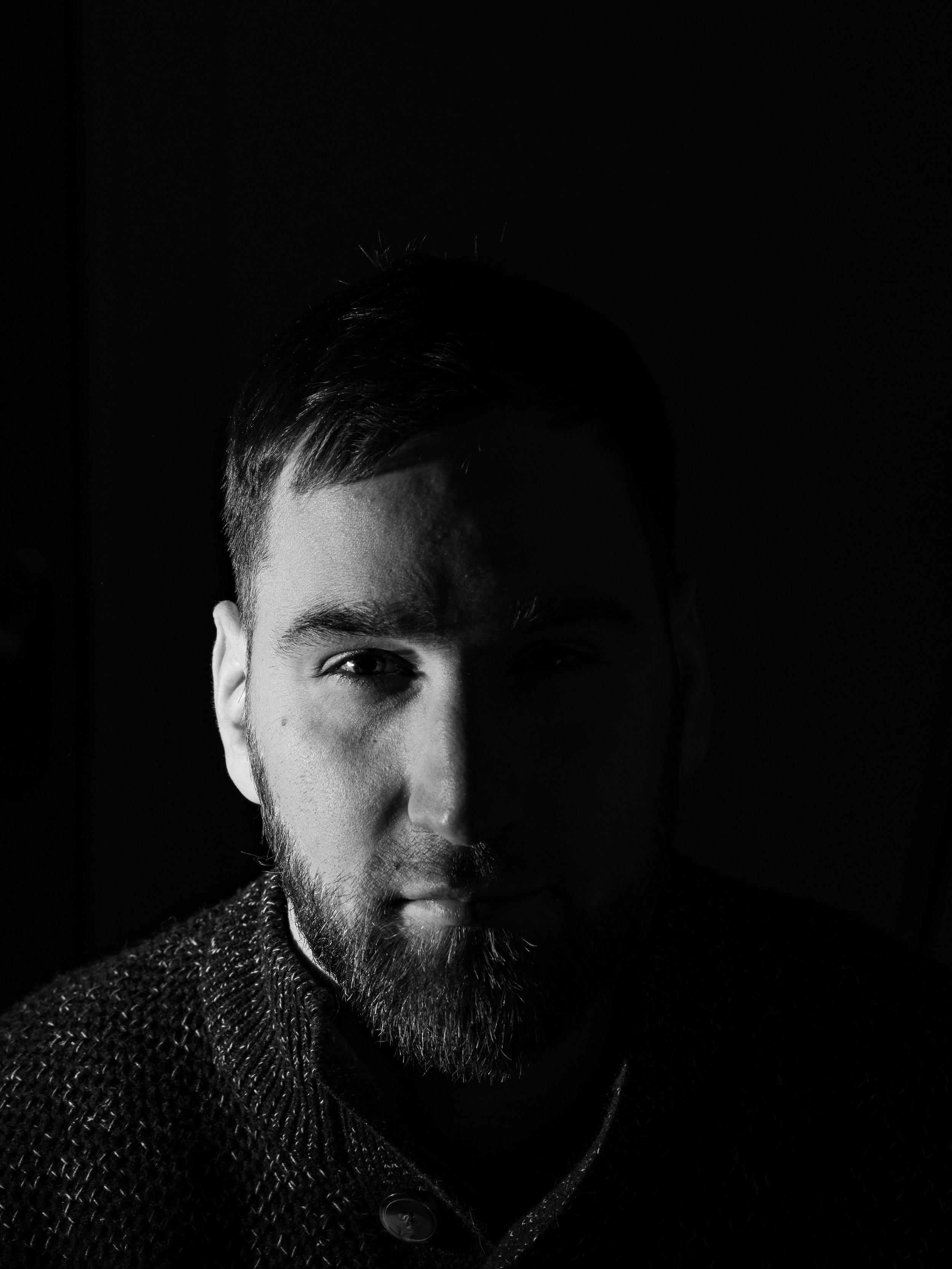 Black and white photo of a young man with a beard, one side of his face illuminated, half of his face in shadow, with dark background.