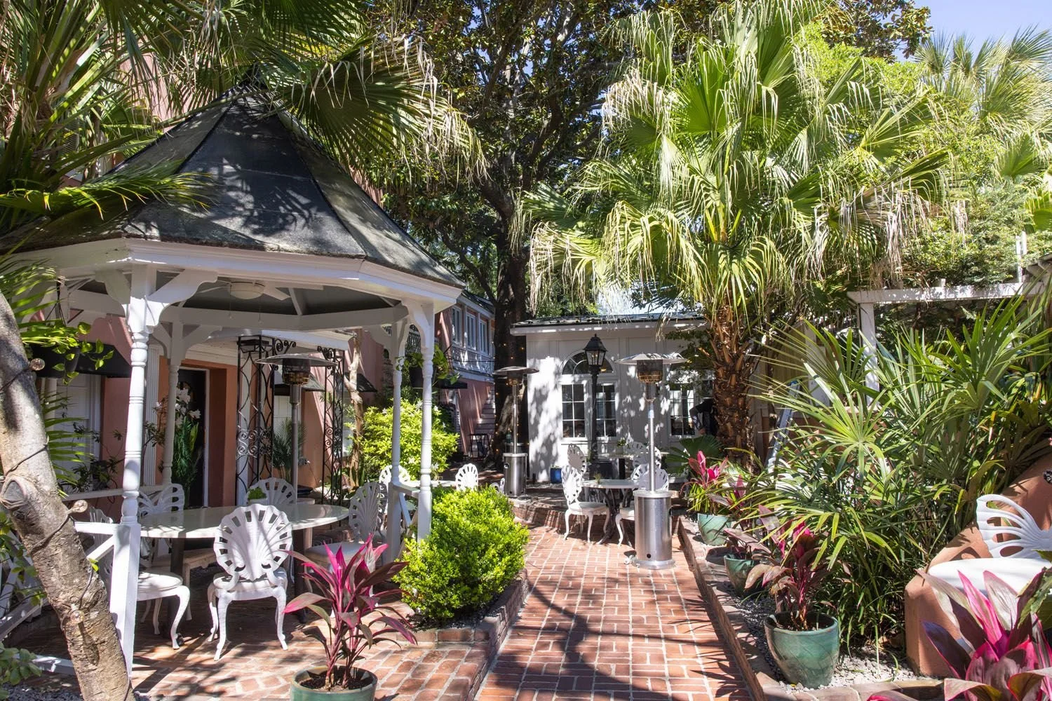 Outdoor patio area with white furniture, potted plants, trees, and a brick pathway.