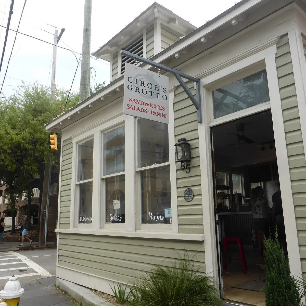 Exterior of a small sandwich shop called Circe's Grotto with a sign advertising sandwiches, salads, and panini. The sidewalk with a crosswalk and a fire hydrant is visible in front.
