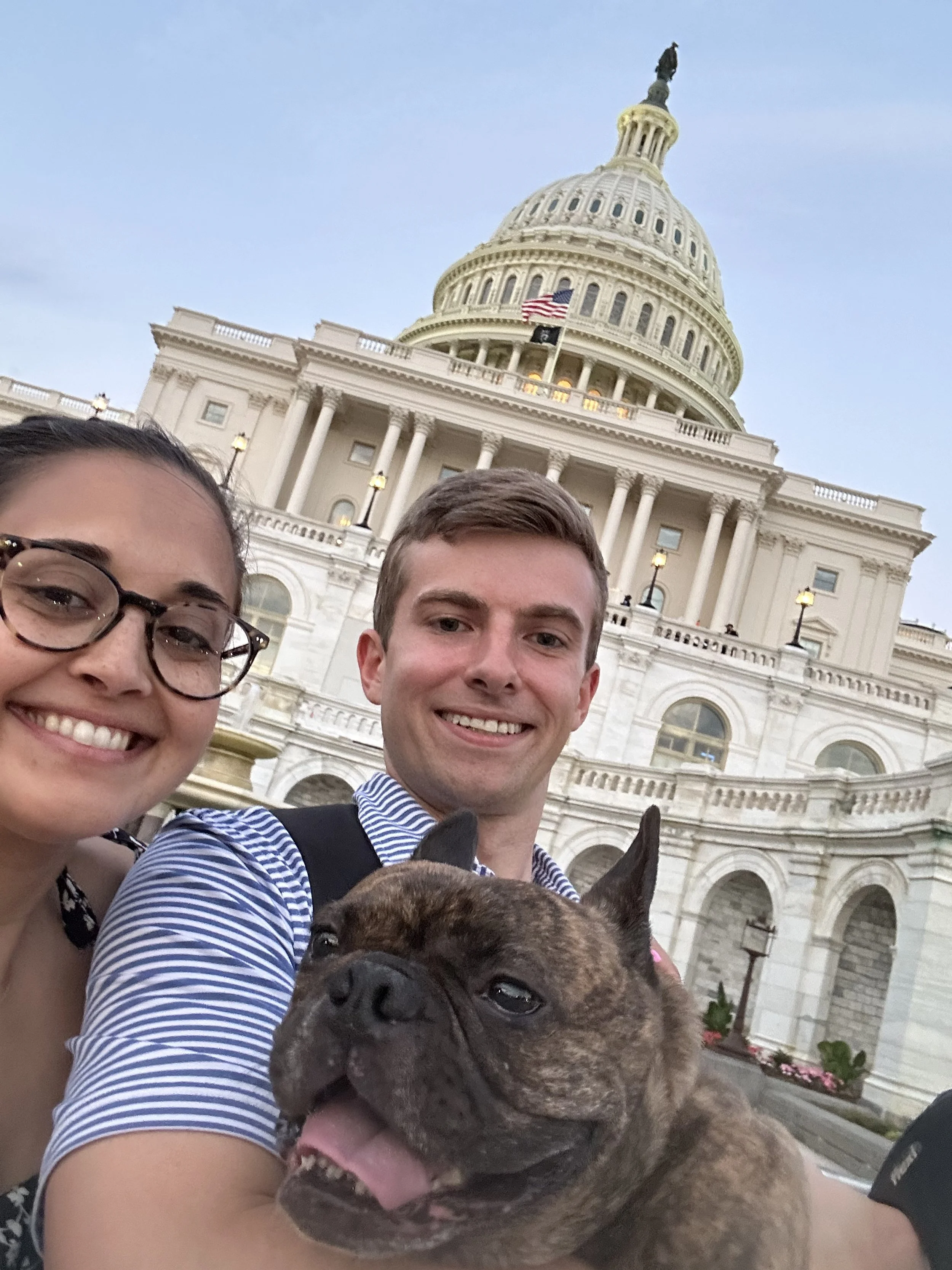 Two smiling people and a dog taking a selfie in front of the U.S. Capitol building.