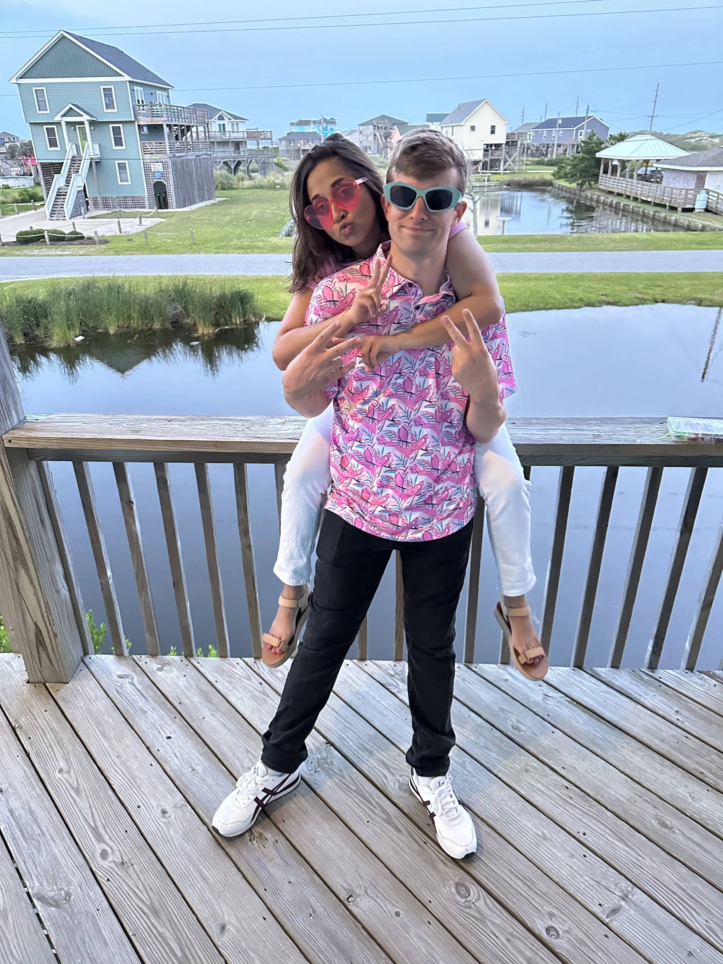 A young man stands on a wooden deck, giving peace signs with both hands, wearing sunglasses and a pink floral shirt. A young girl is piggybacking on him, wearing pink sunglasses, a sleeveless top, white pants, and sandals, making a kissing face. They