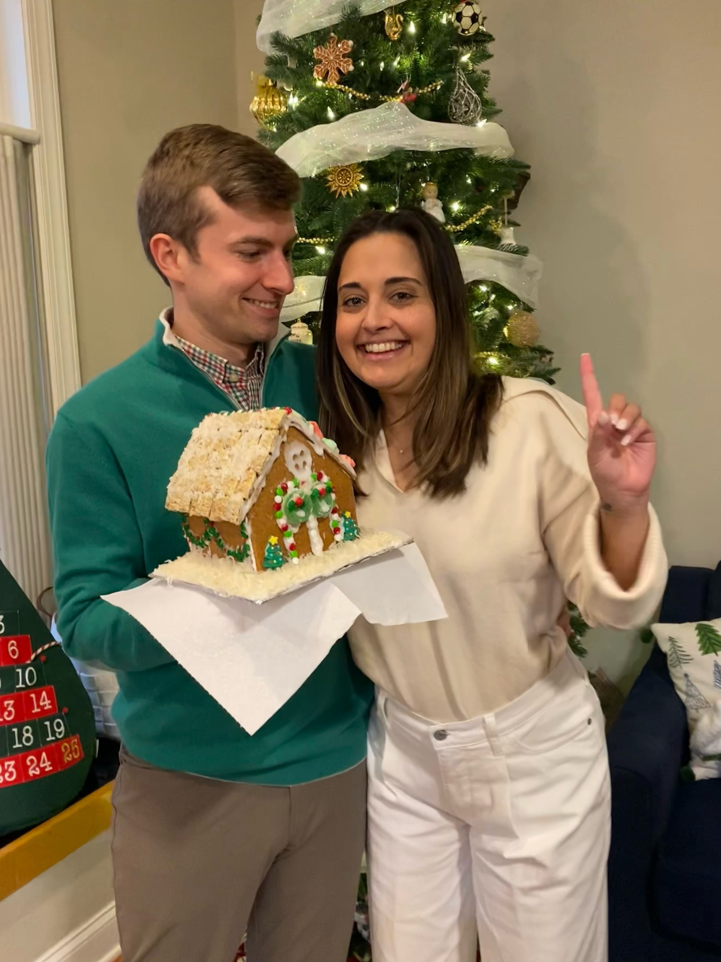 A man and woman smiling and posing with a gingerbread house in front of a decorated Christmas tree.