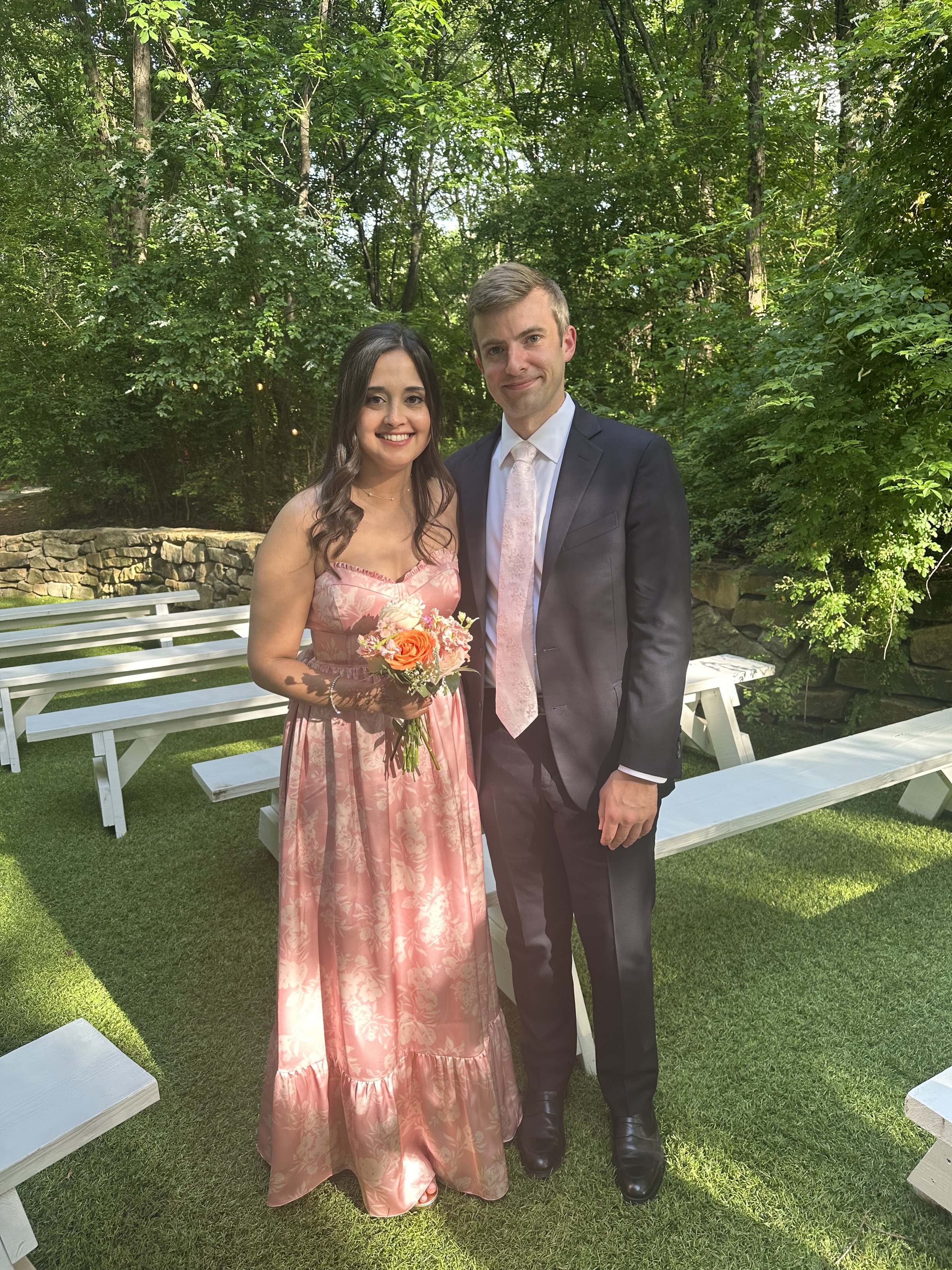 A woman in a pink floral dress holding a bouquet of flowers and a man in a black suit standing outdoors in a garden with green trees and white benches.