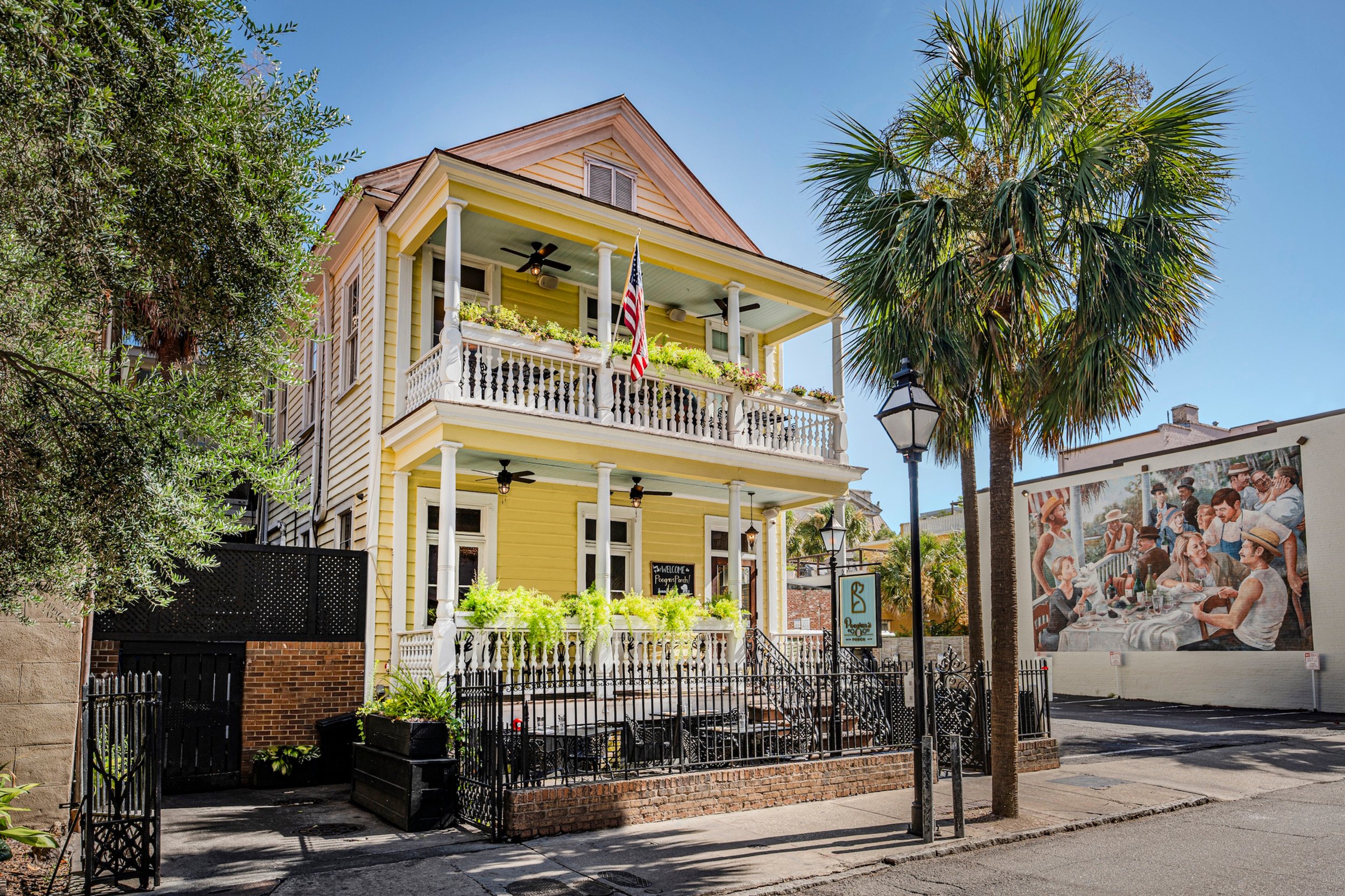 Yellow three-story house with a wraparound porch, balcony, and potted plants, surrounded by a black fence, with palm trees and street lamps, and a mural on the adjacent wall depicting a group of people dining outdoors.
