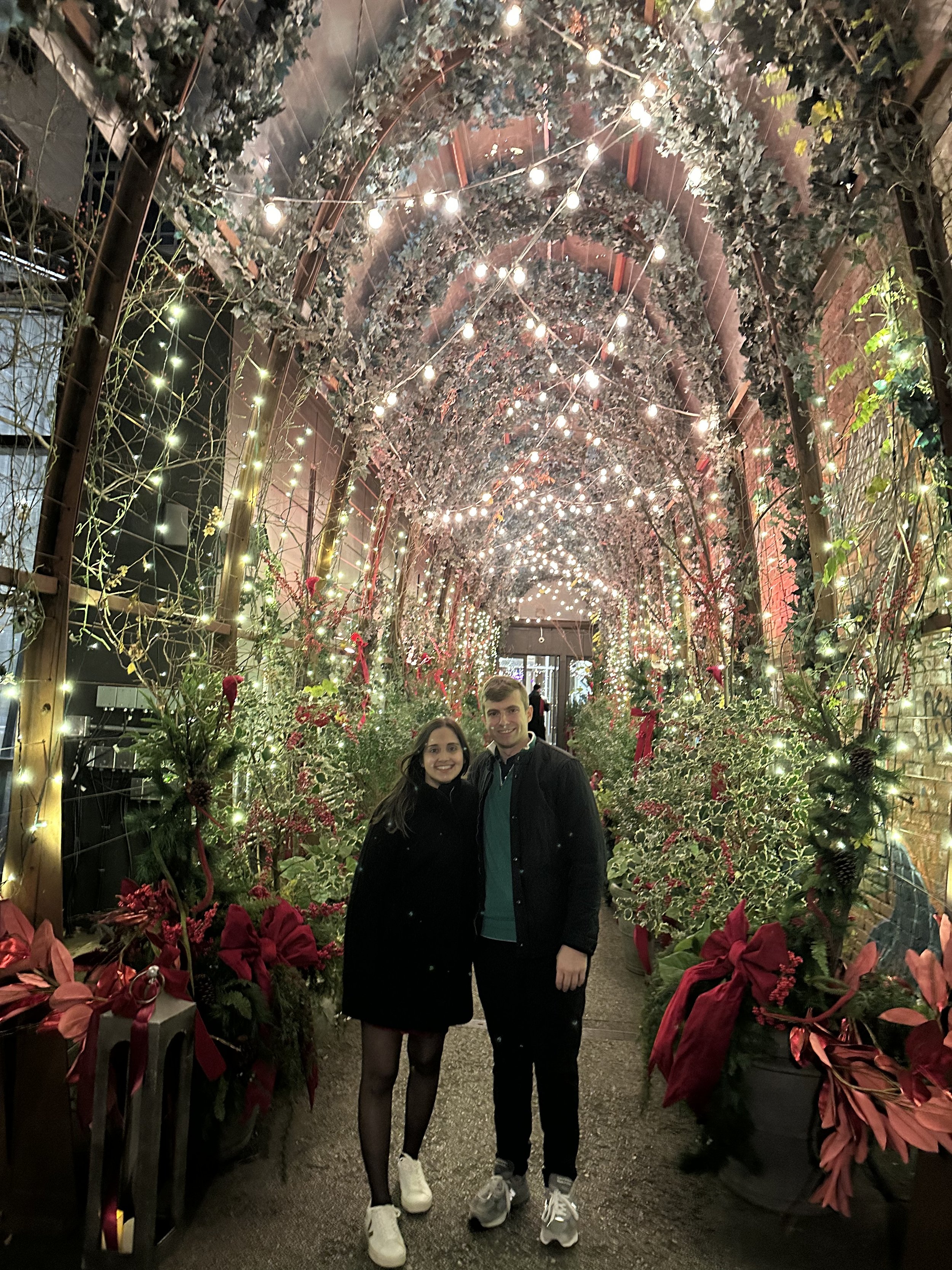 A smiling young couple standing in a brightly decorated holiday walkway with string lights, greenery, and red flowers surrounding them.
