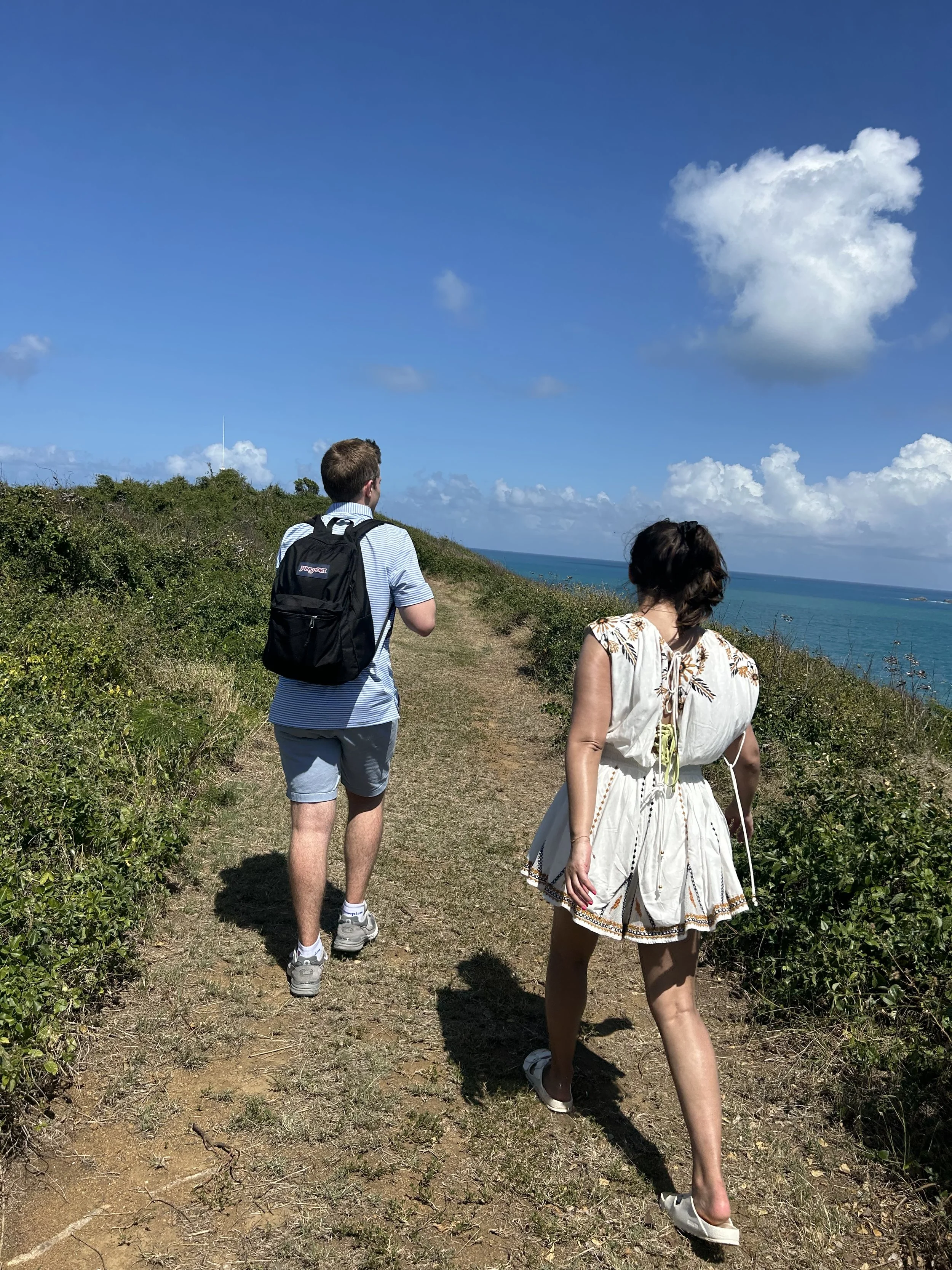 A man and woman walking on a grassy coastal trail with the ocean visible to the right, blue sky, and white clouds overhead.