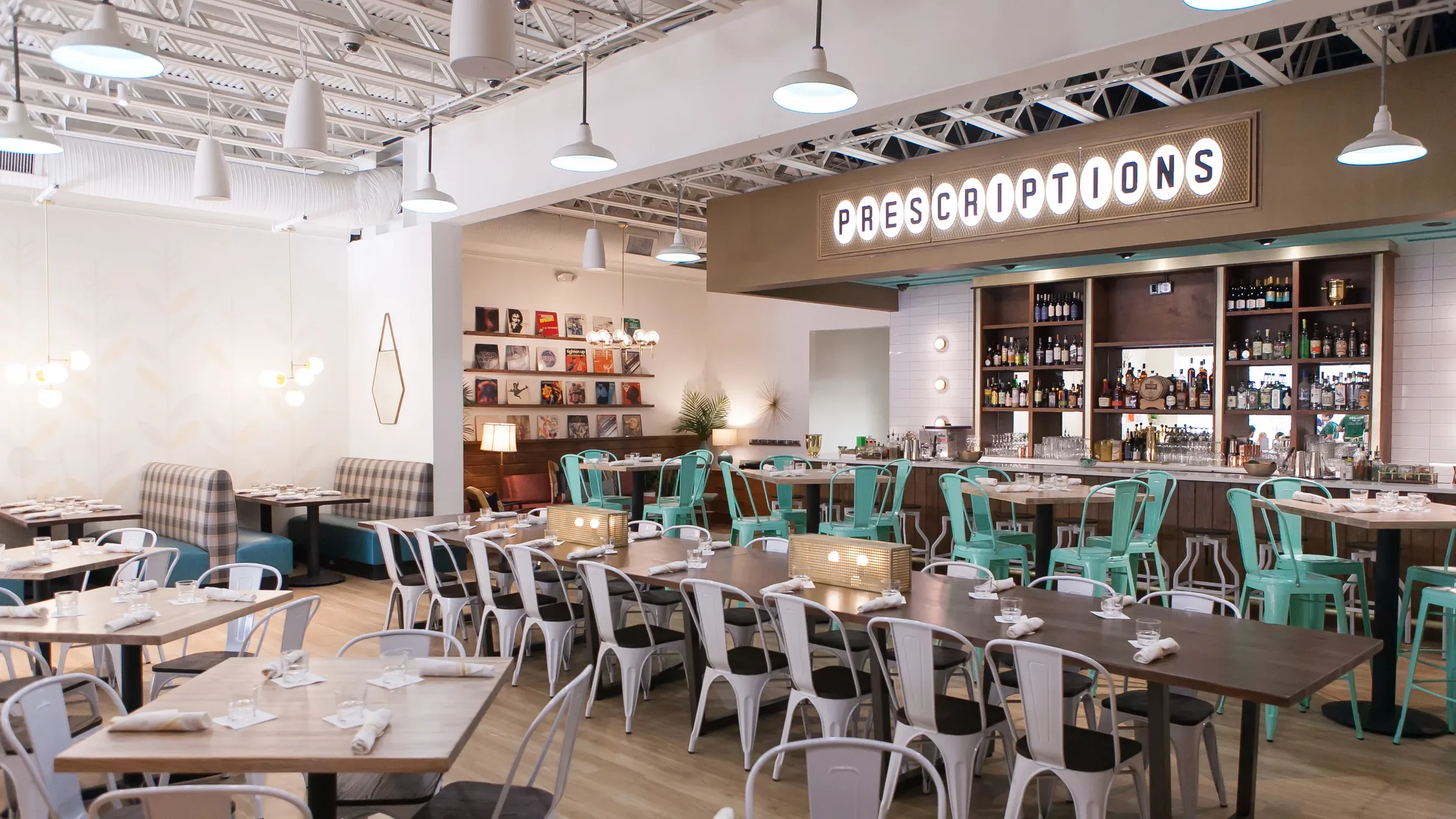 Interior of a restaurant bar with tables set for dining, a bar area with bottles of alcohol, and a sign reading 'Prescriptions' hanging from the ceiling.