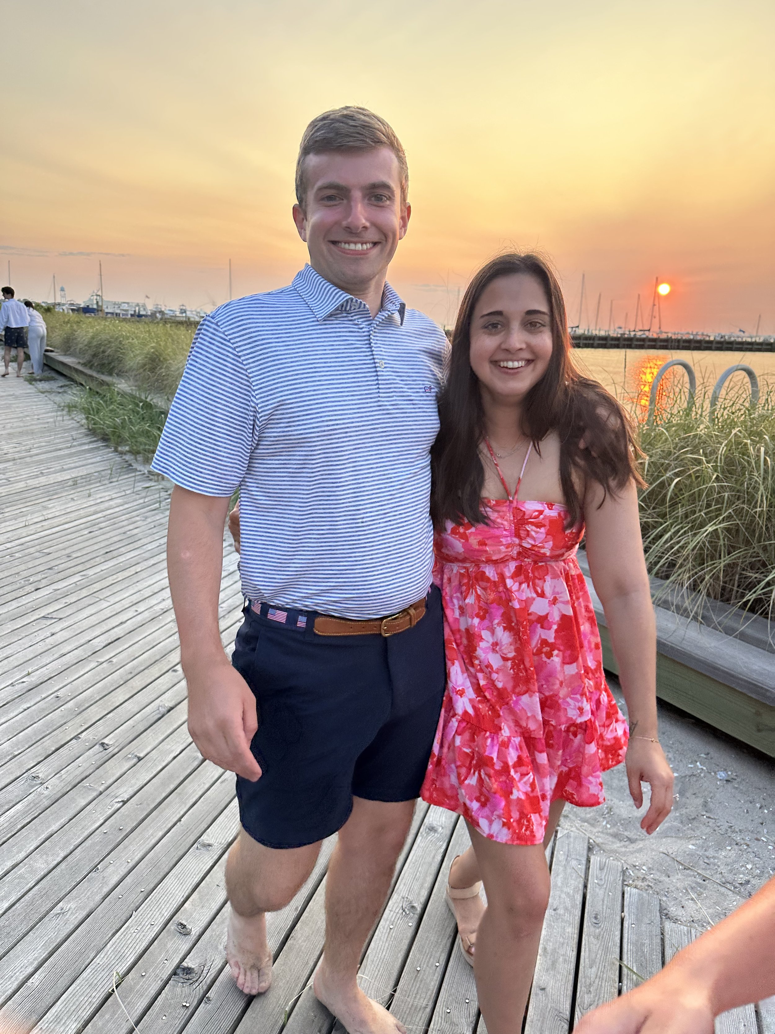 A young man and woman standing barefoot on a wooden dock during sunset, smiling at the camera with a marina and sailboats in the background.