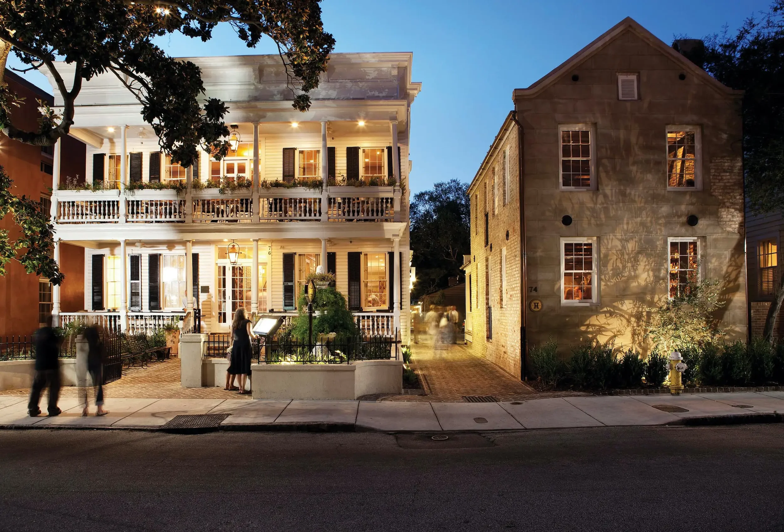 A three-story, white, historic Southern-style house with a wrap-around porch, illuminated at dusk, with several people standing on the sidewalk in front. Next to it is a two-story brick building with lit windows, surrounded by trees and plants.