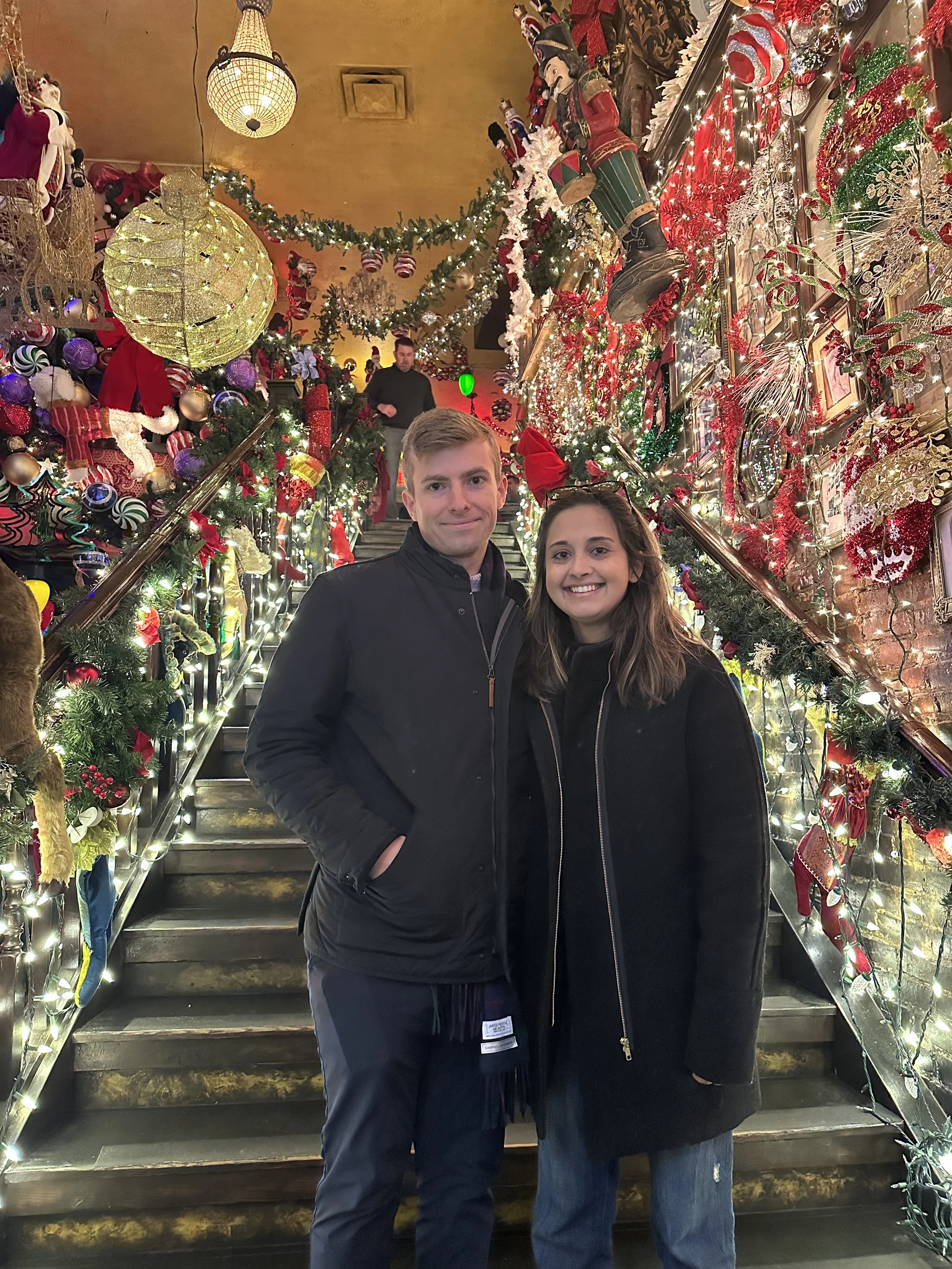 A young man and woman standing closely together on a festive staircase decorated for Christmas, with ornaments, lights, stockings, and nutcracker figures in a decorated indoor setting.