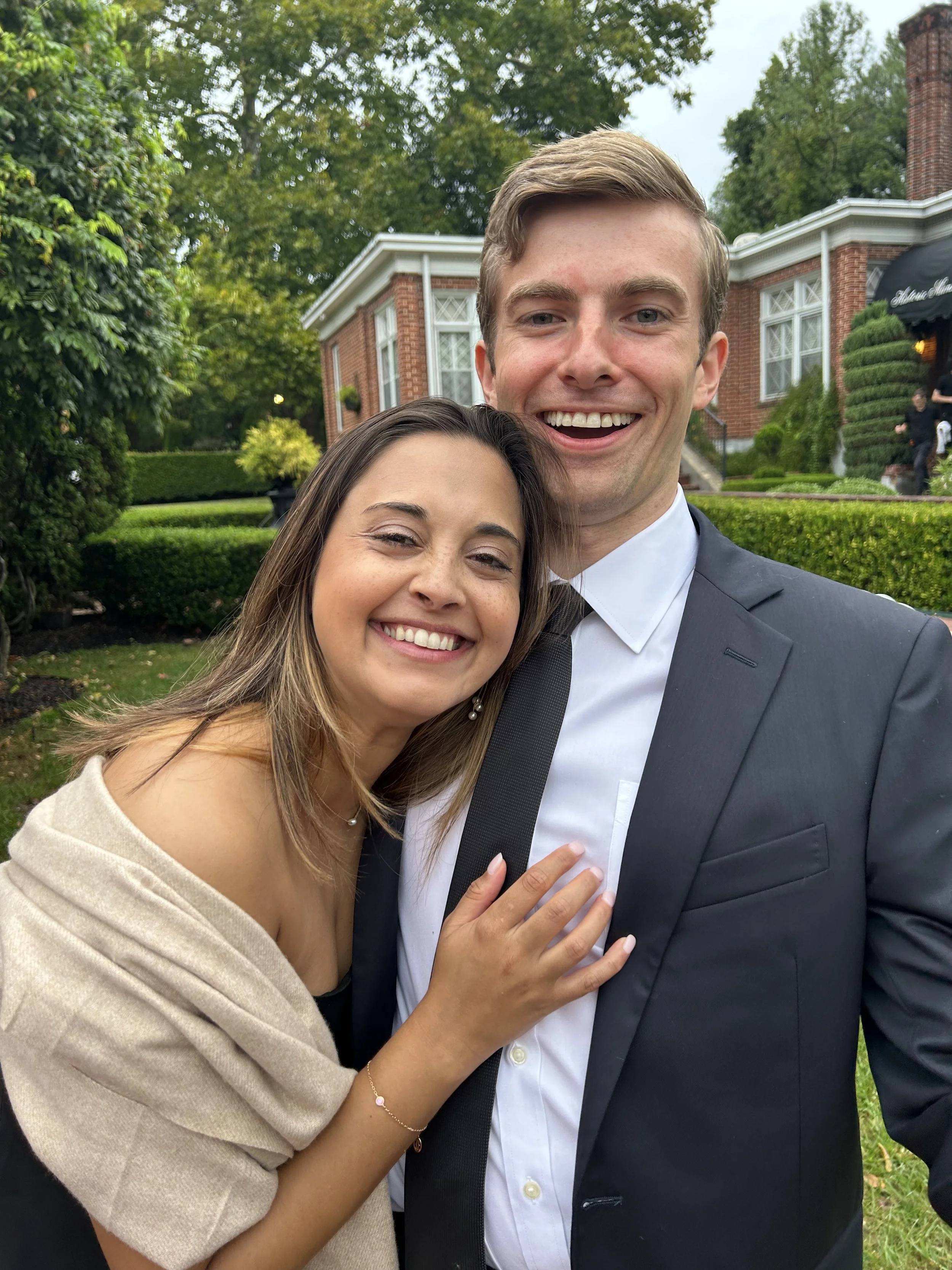 A smiling young woman and young man in formal attire taking a selfie outdoors with a large brick house and lush green trees in the background.