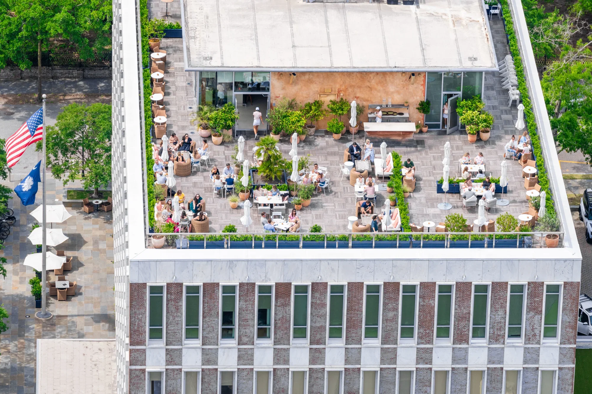 Aerial view of a rooftop patio with people sitting at tables, lounging, and walking. The patio is decorated with potted plants and has a mix of outdoor furniture, including umbrellas and chairs. Surrounding the building are trees and a city street with American and South Carolina flags.