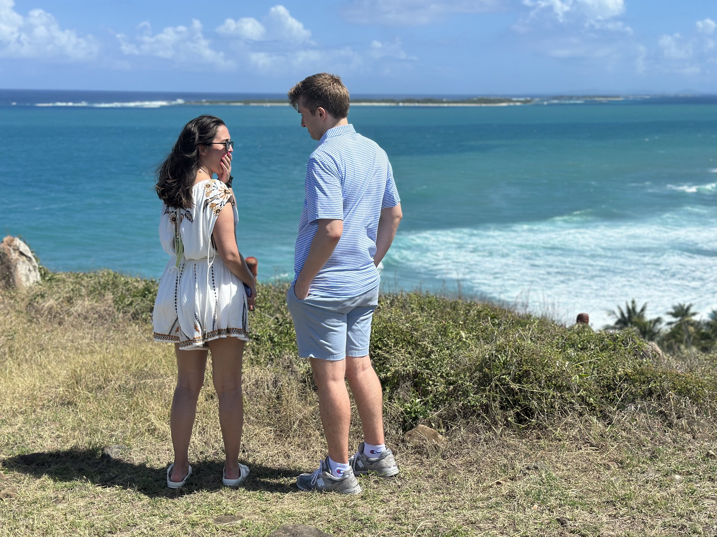 A man and woman standing on a grassy hill near the ocean with waves and a distant shoreline, engaged in conversation on a sunny day.