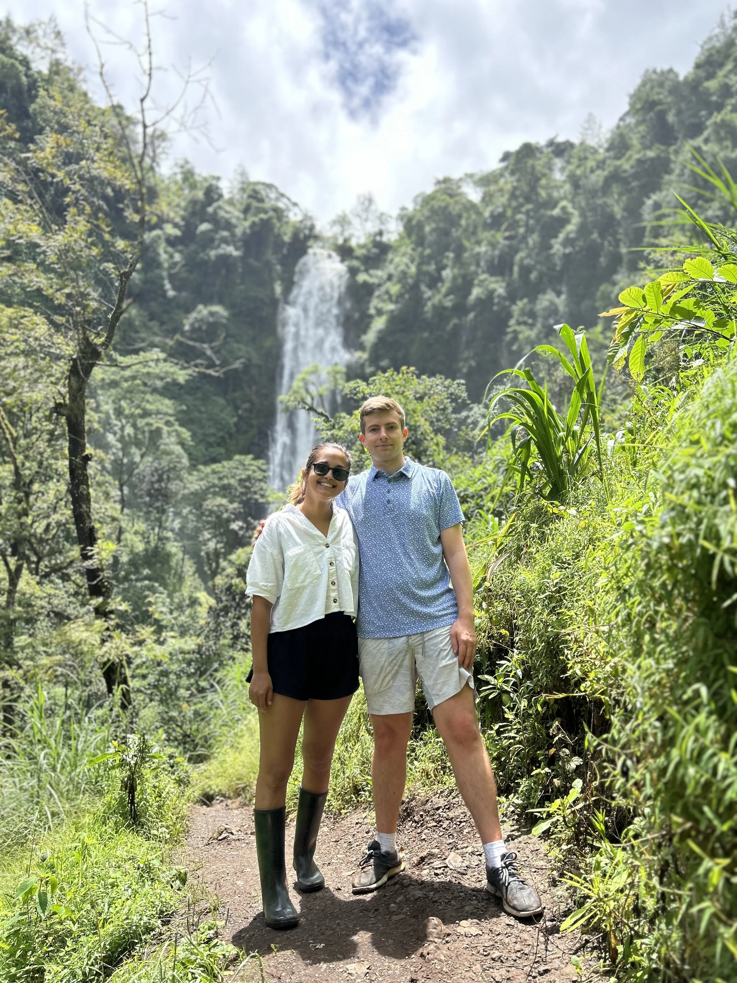 A couple standing on a forest trail with a waterfall in the background, surrounded by lush greenery and trees.