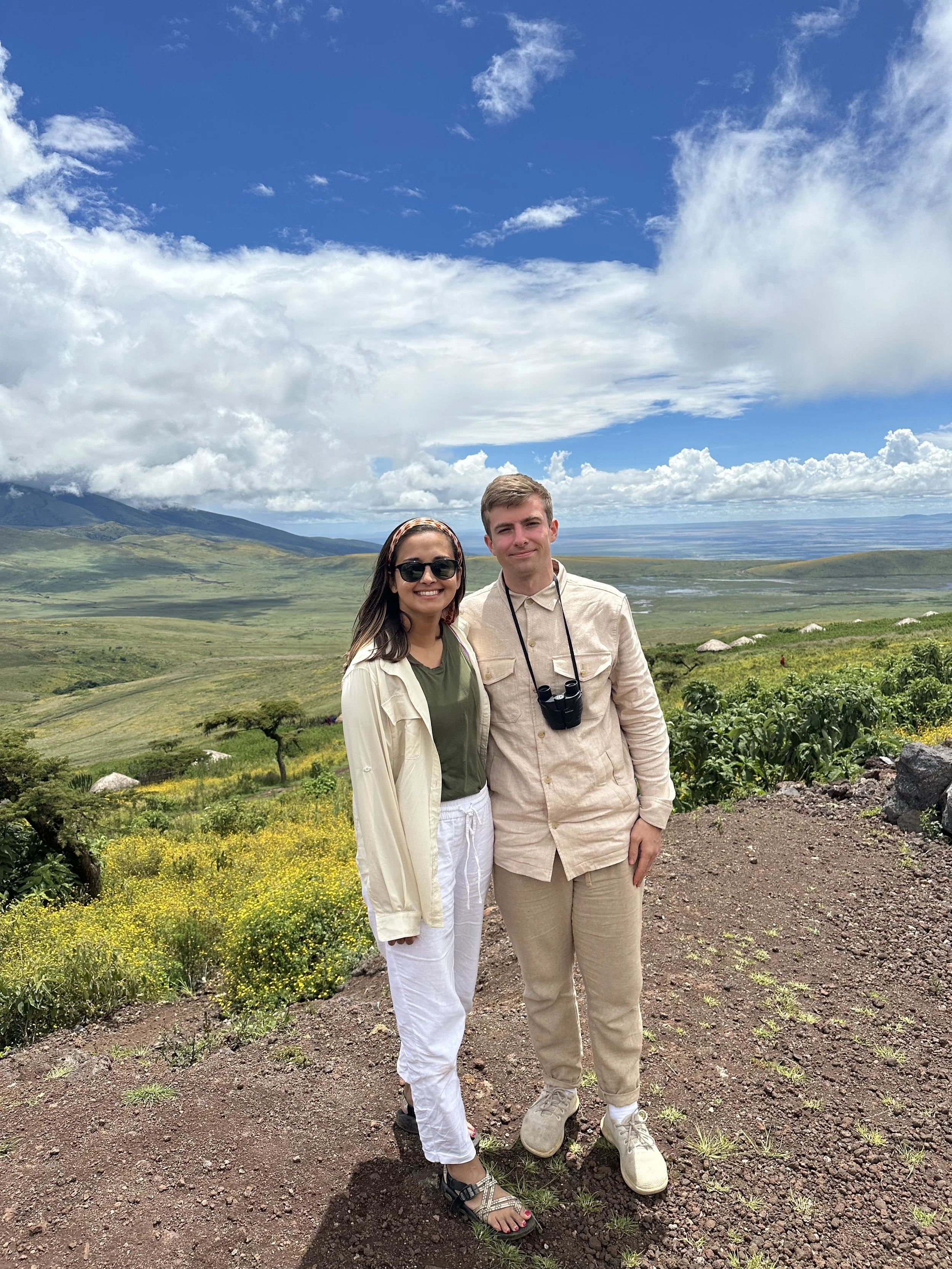 Two people standing on a hillside with green fields and mountains in the background under a partly cloudy sky.