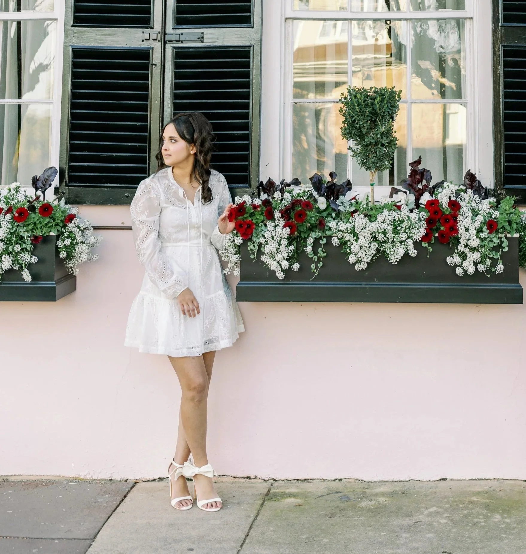 A young woman in a white dress and white high-heeled shoes with a bow stands in front of a pink wall with flower boxes below a black window with shutters. She is holding a red flower and looking to her left, surrounded by greenery and white and red flowers.