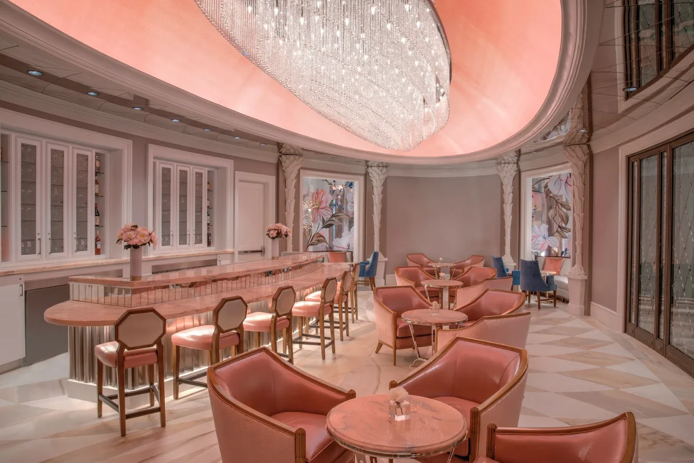 Elegant lounge bar with pink and blue armchairs, marble tables, a pink marble bar counter, floral decor, and a large crystal chandelier overhead.