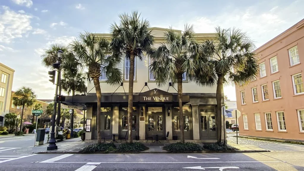 A restaurant named 'The Vendue' with palm trees in front, located on a city street corner with pastel-colored buildings and a traffic light.
