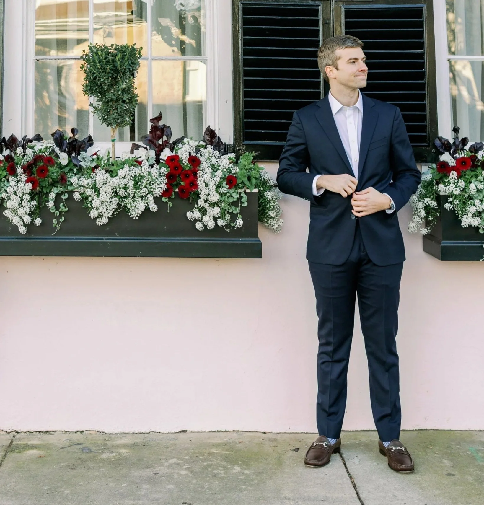 A man in a navy suit standing outdoors in front of a wall with window flower boxes filled with white baby's breath and red flowers, near black shutters.