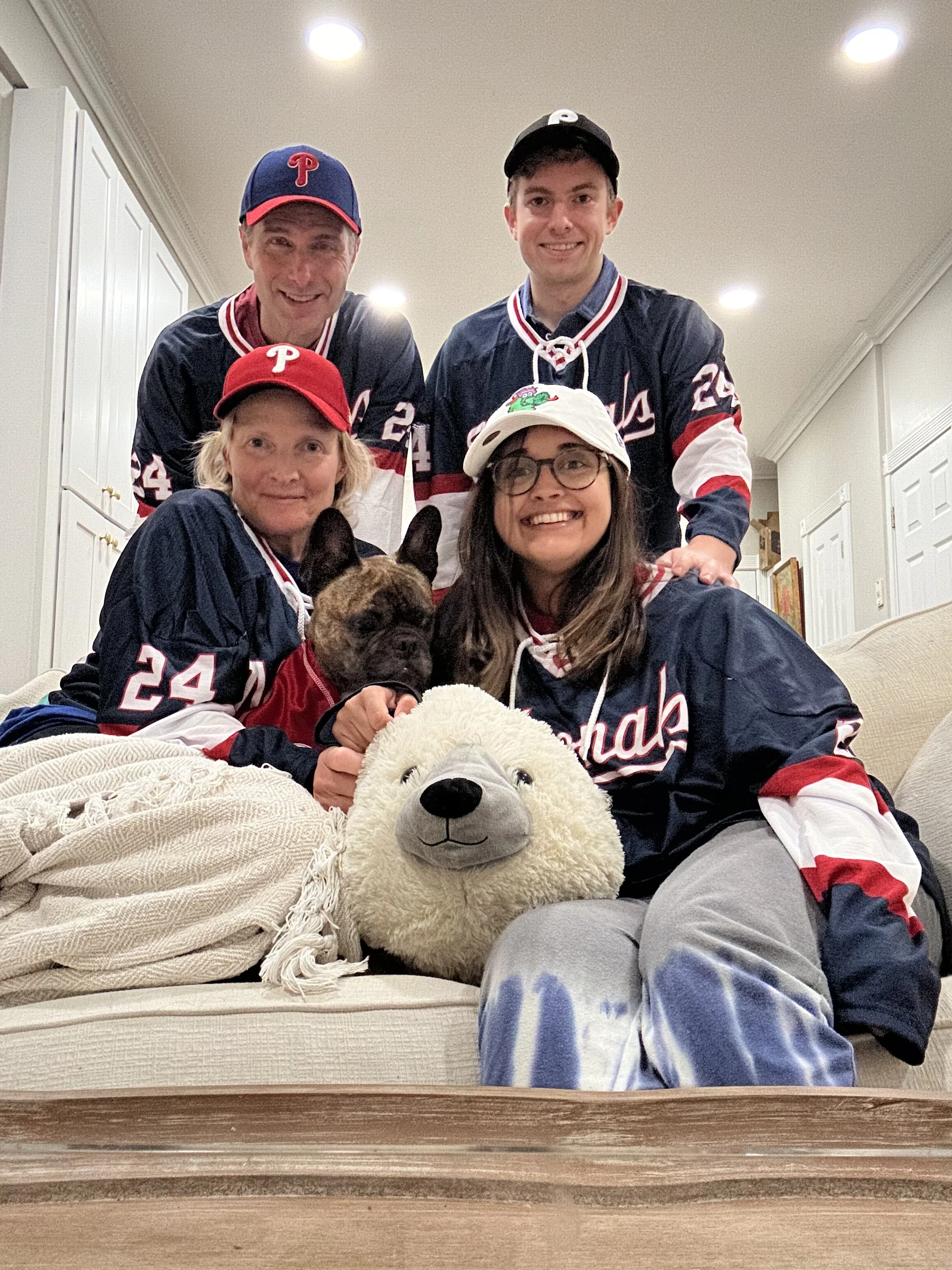 A group of five people in sports jerseys and caps, accompanied by a dog and plush toys, gathered together in a living room for a photo.