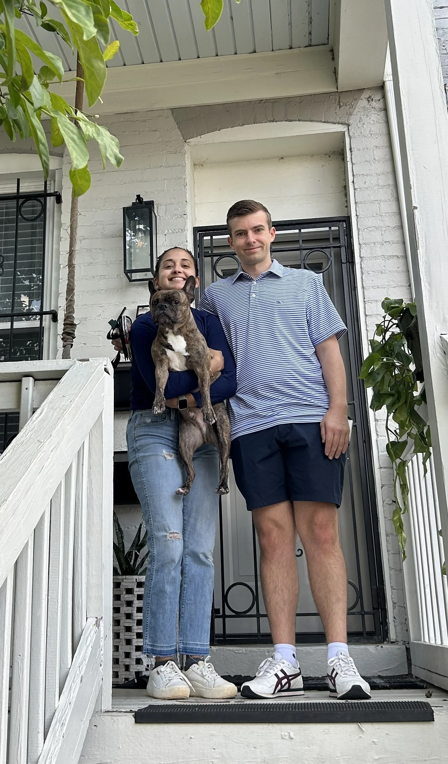A smiling woman holding a small dog and a man standing next to her on a front porch with white brick walls and black metal gate.