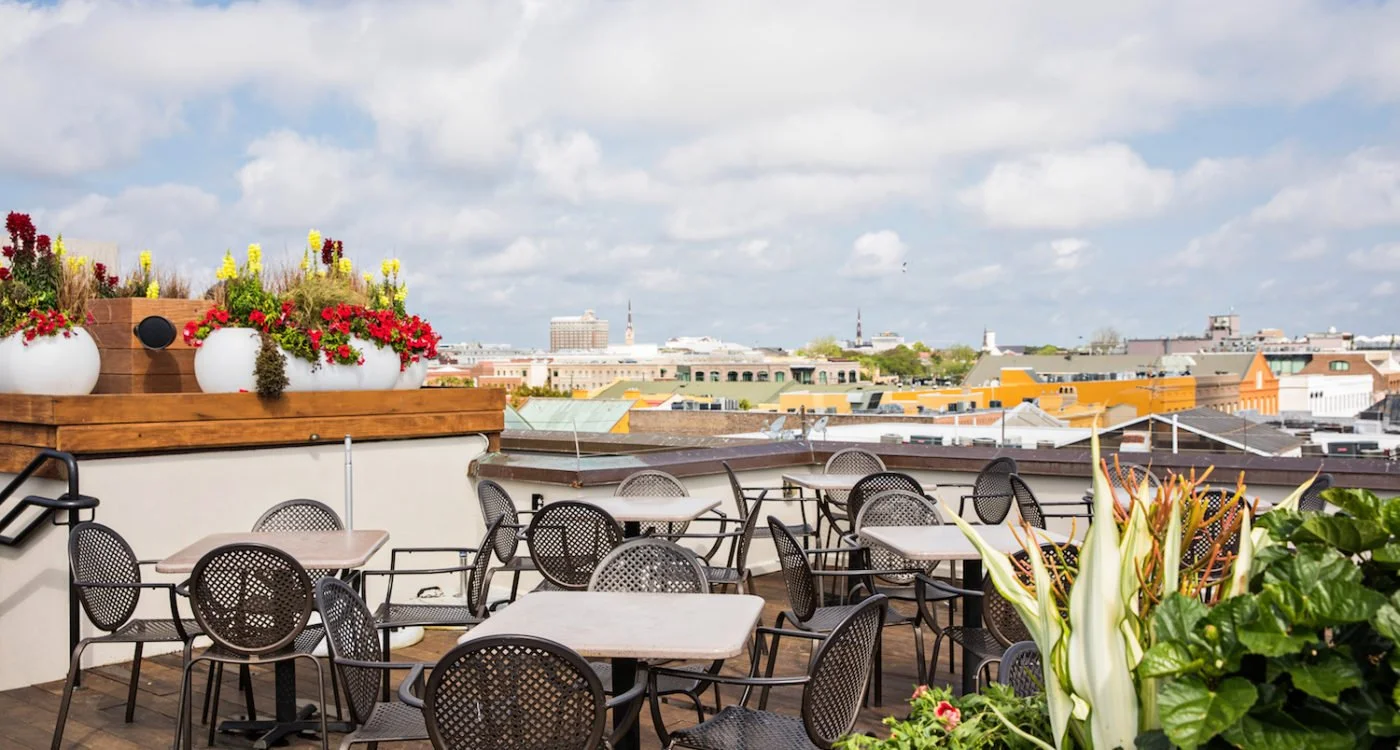 Rooftop patio with black metal tables and chairs, potted plants, and city skyline in the background under cloudy sky.
