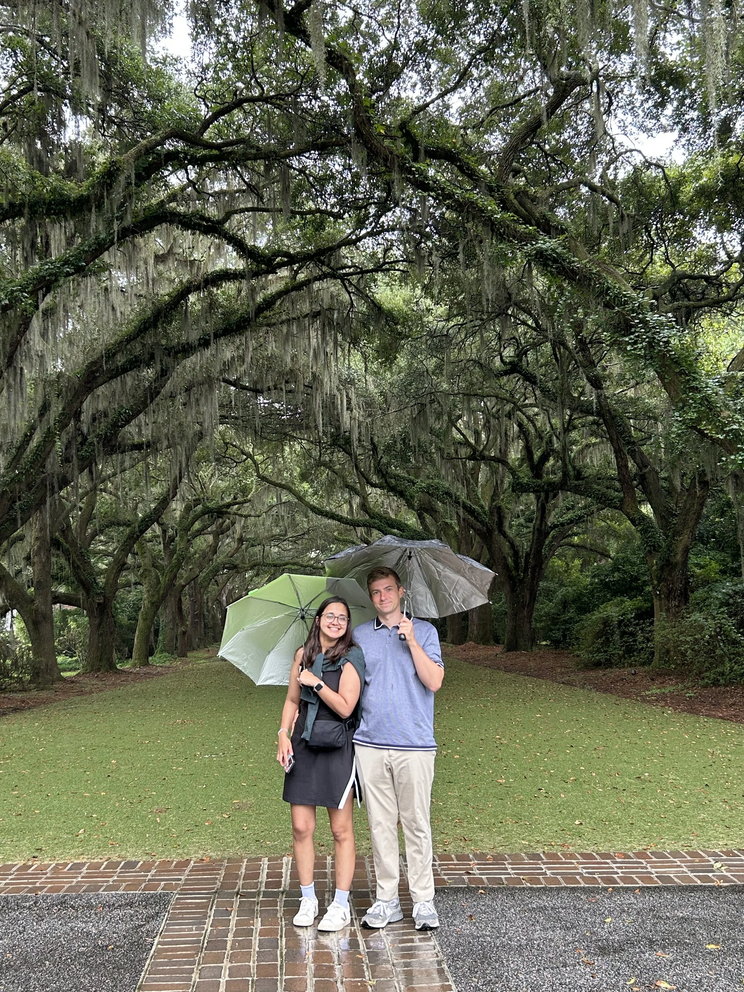 A young couple stands on a brick pathway under umbrellas in a lush, green park with large, hanging moss-covered trees in rainy weather.
