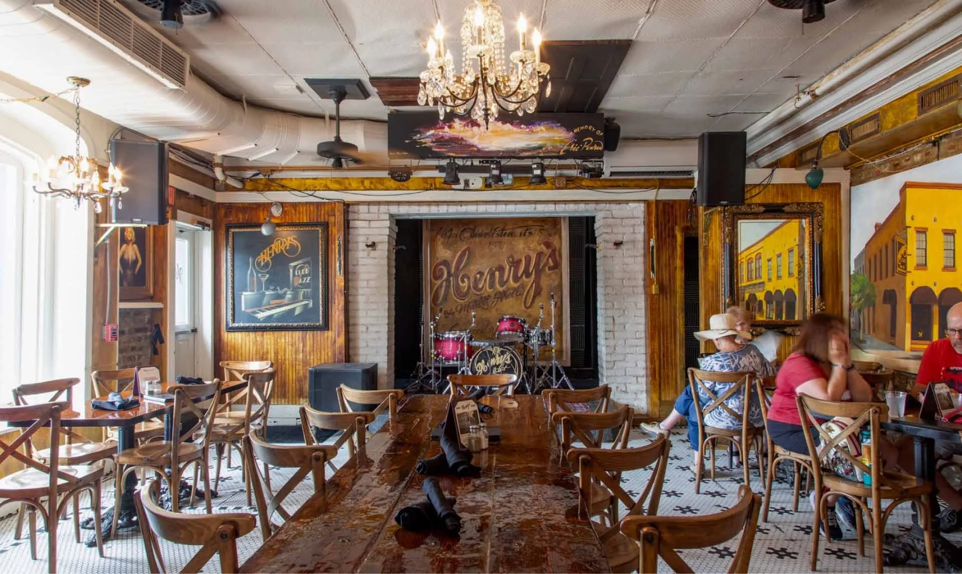 Interior of a lively bar with people sitting at tables and the counter. Neon signs and decorations adorn the walls, including beer brands, skull artwork, and vintage posters. There is a staircase leading to an upper level with more signage, lamps, and hanging decorations.