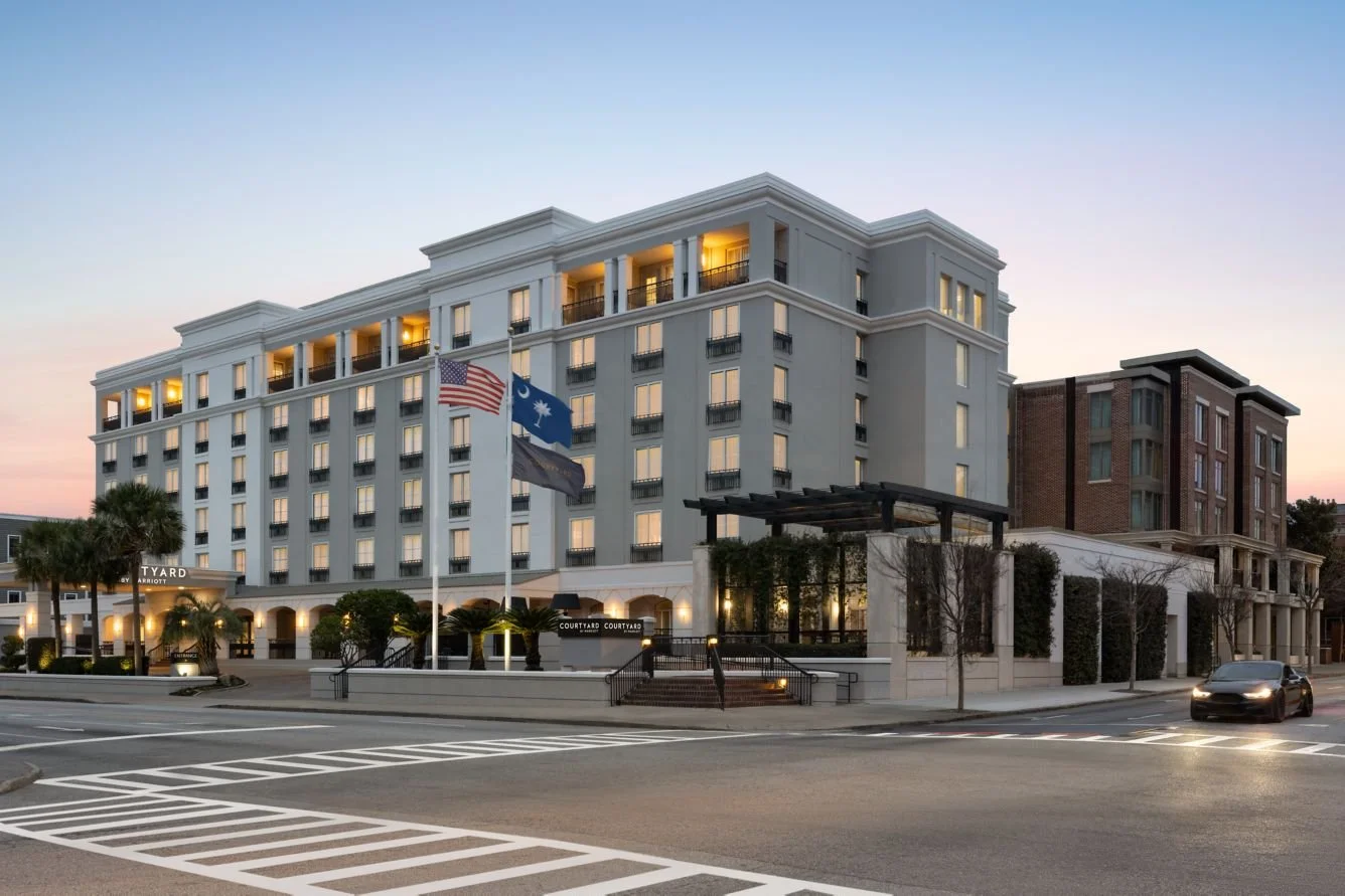 A modern hotel building at sunset with flags of the United States and other countries flying in front. The hotel has multiple floors with lit-up windows and a landscaped entrance.