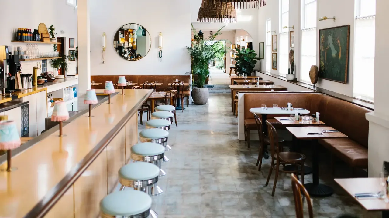 Empty restaurant interior with bar seating and booths, featuring wooden furniture, large windows, art on the walls, and potted plants.