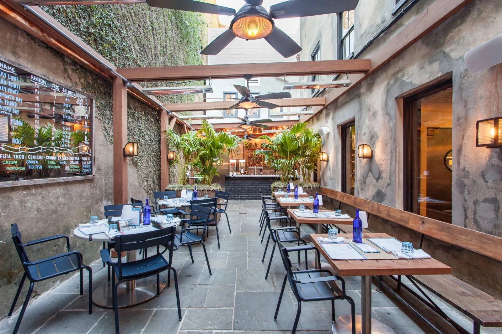 Outdoor restaurant patio with wooden tables, black chairs, blue bottles, and place settings, surrounded by green plants, with ceiling fans and wall-mounted lights, and a chalkboard menu on one wall.