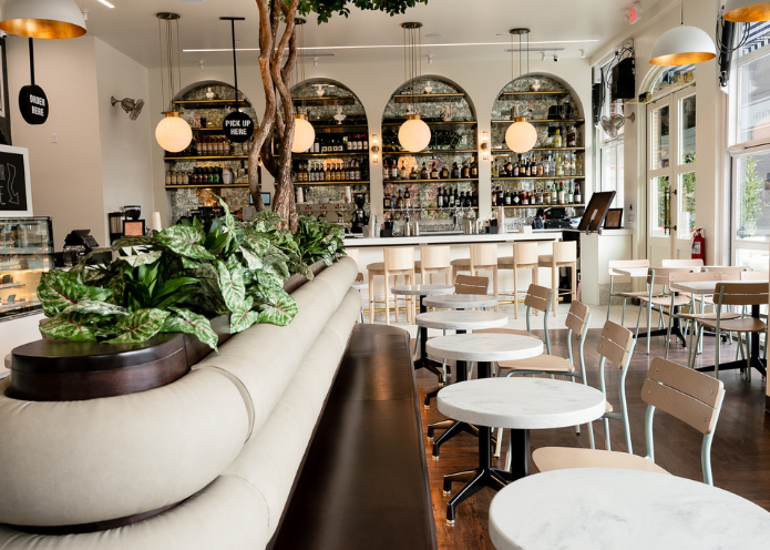 Interior of a modern cafe with white marble tables and beige chairs, a cozy seating area with a beige sofa decorated with green plants, and a bar with shelves stocked with bottles and glasses in the background, illuminated by pendant lights.