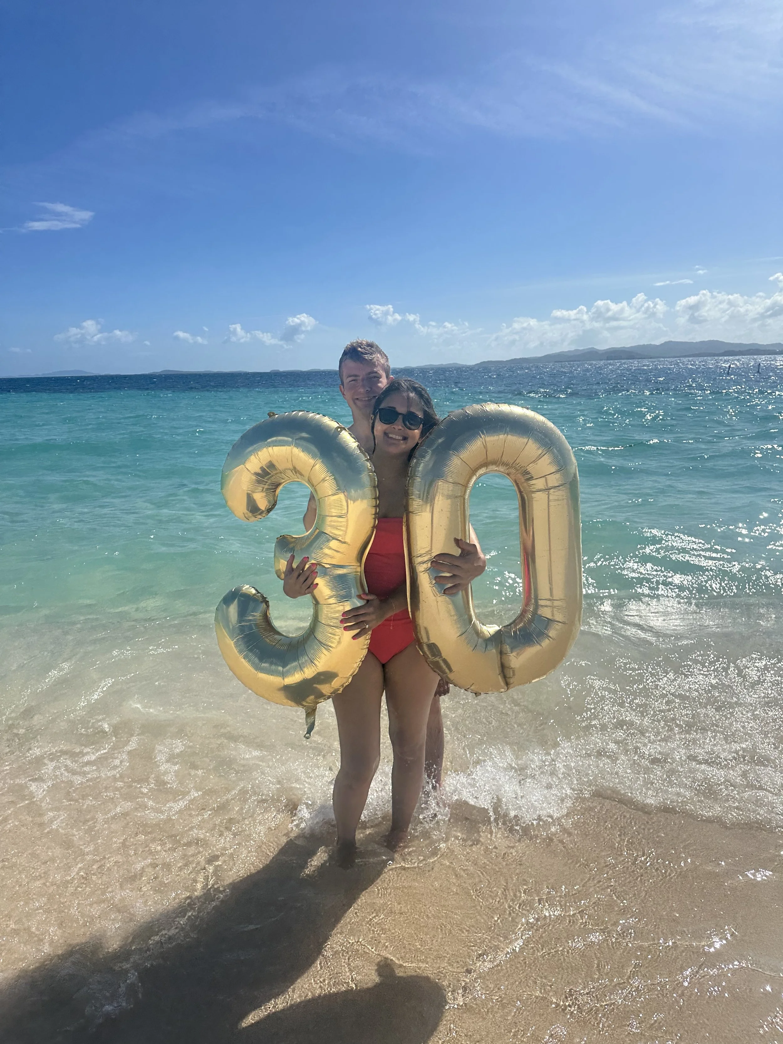 Smiling woman in red swimsuit and sunglasses holding gold inflatable balloons shaped as the number '30' while standing in the surf at the beach, with a man partially visible behind her, and the ocean, blue sky, and distant land in the background.