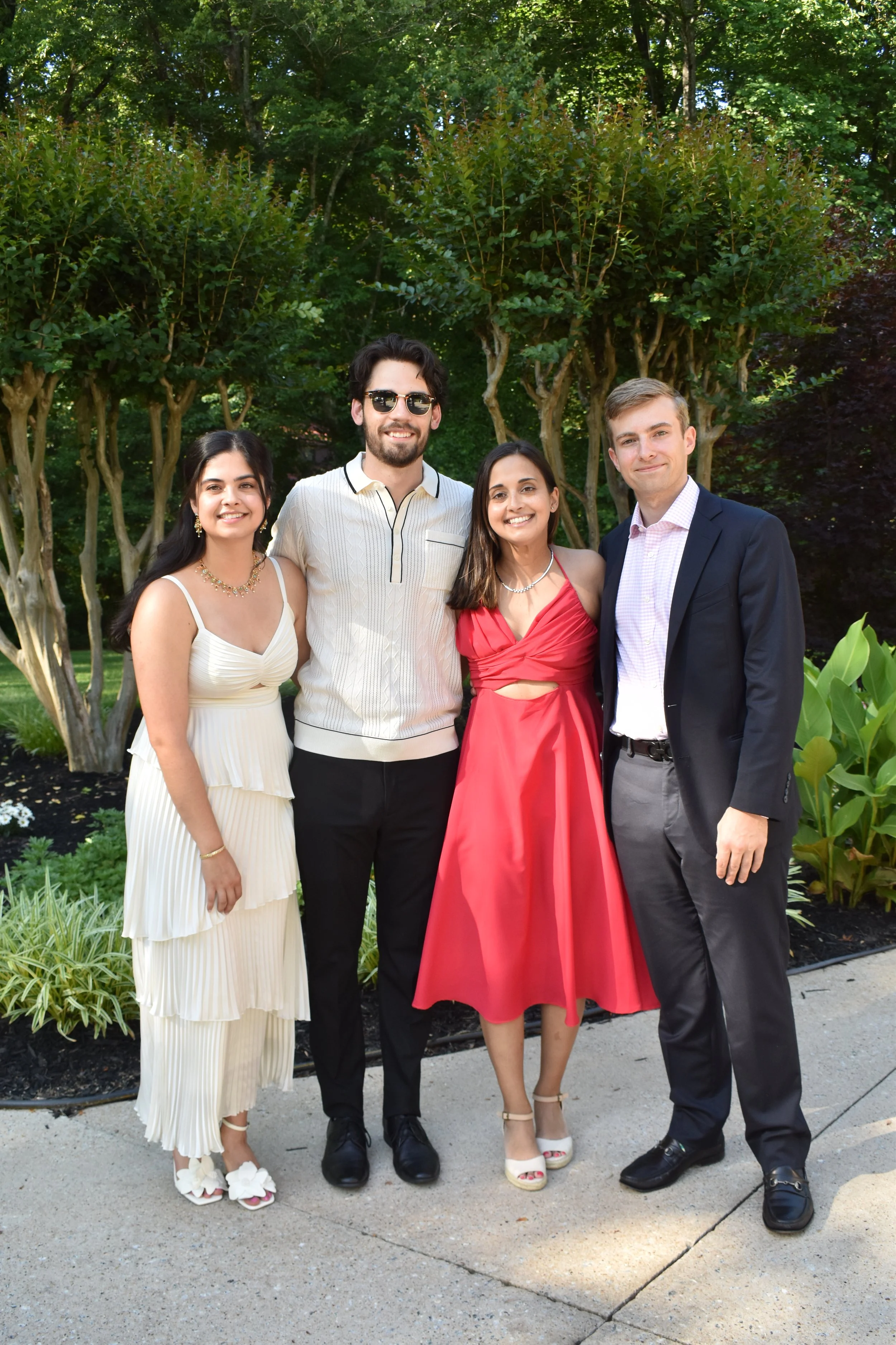 Group of four young adults dressed in formal attire, standing outdoors in front of green trees and plants, smiling at the camera.