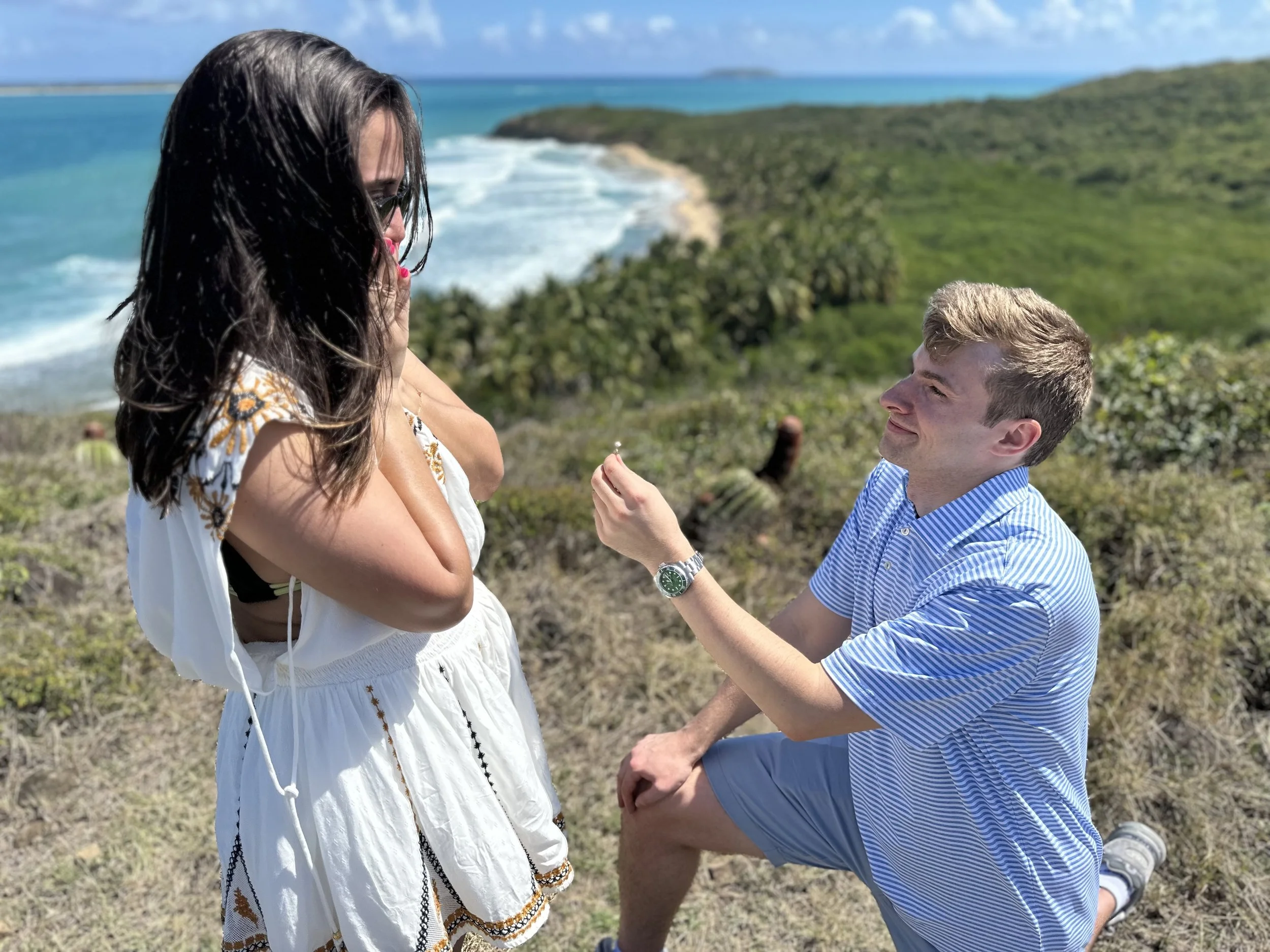 A man proposing to a woman on a scenic coastal hill with ocean and greenery in the background.