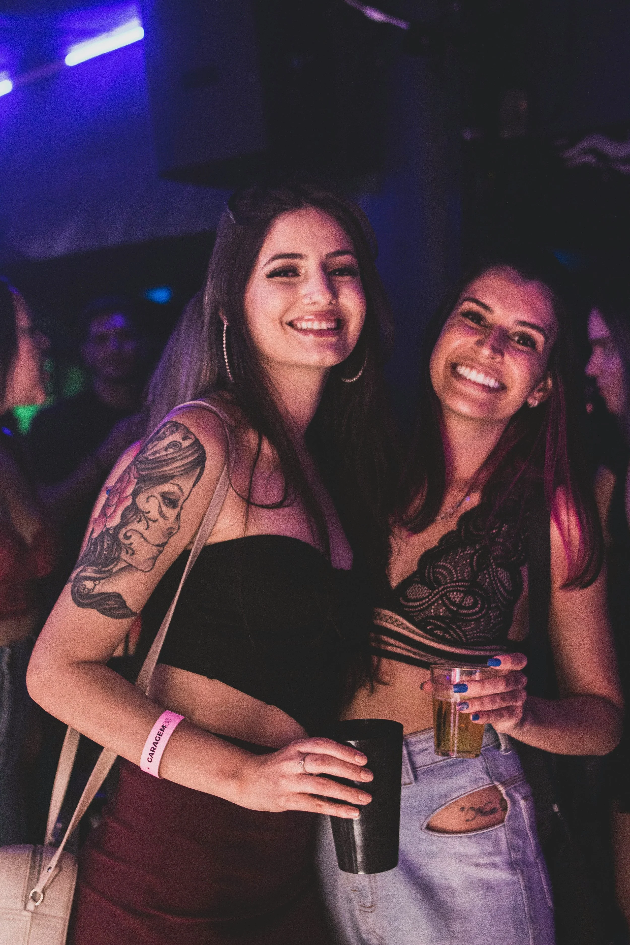 Two young women smiling and holding drinks at a nightclub or party, with colorful lighting in the background.