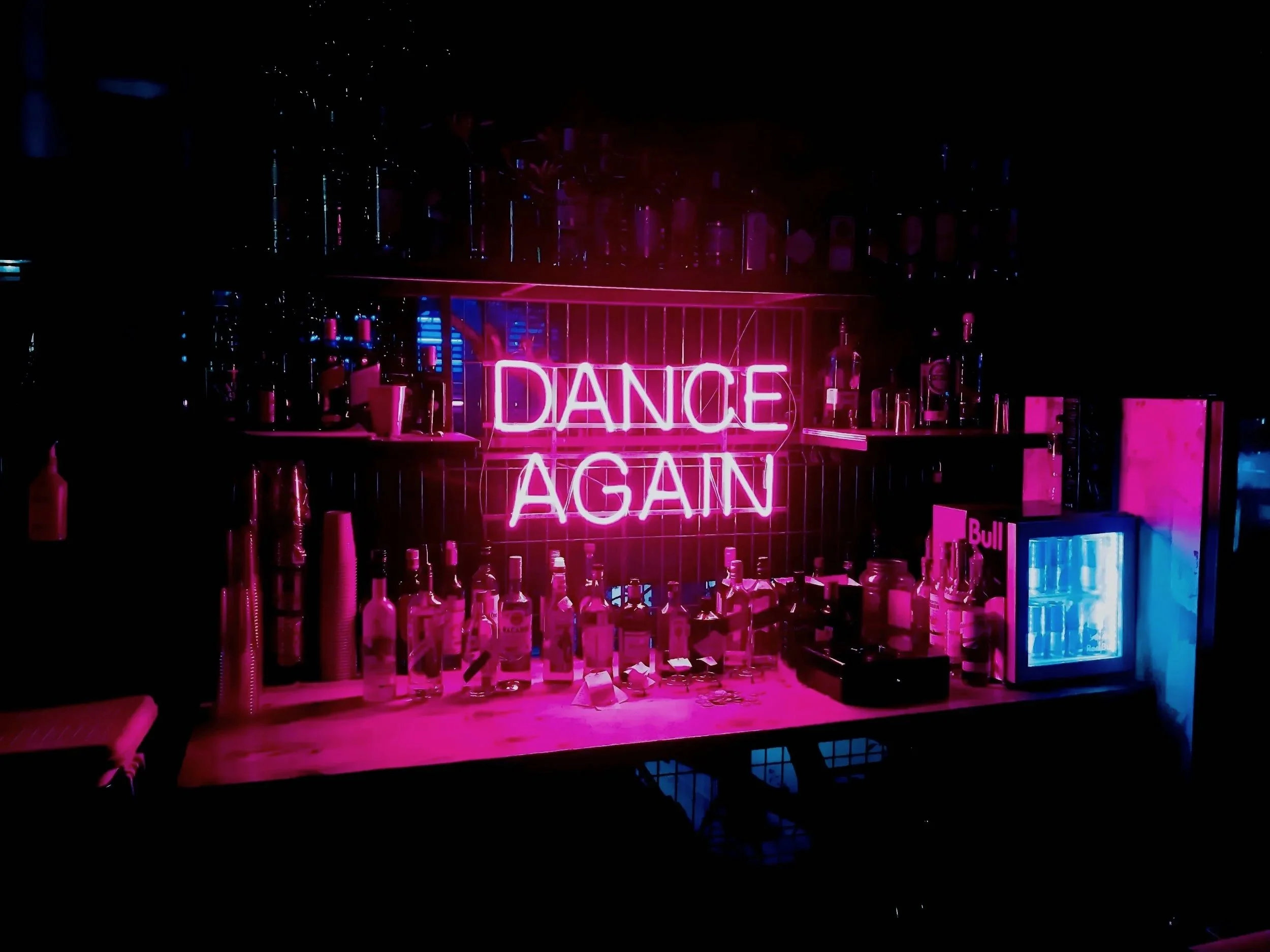 Neon pink sign reading 'Dance Again' hanging above a bar area with bottles and bar equipment illuminated by pink and blue lighting.