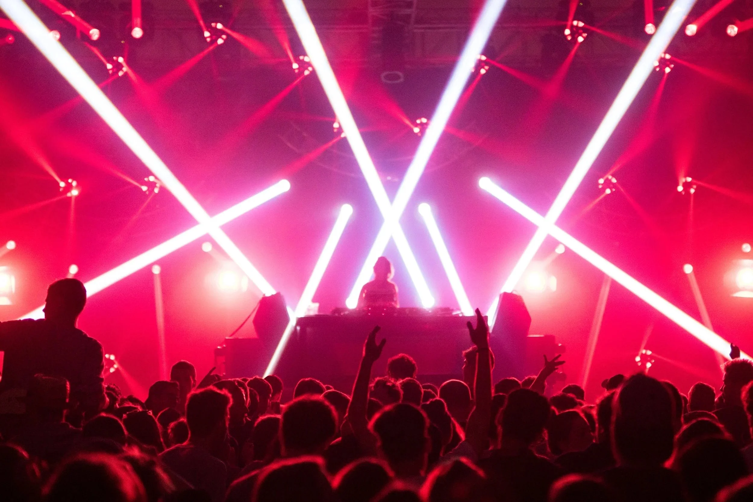A DJ performing on stage with bright pink and white laser lights in a concert with a crowd dancing.