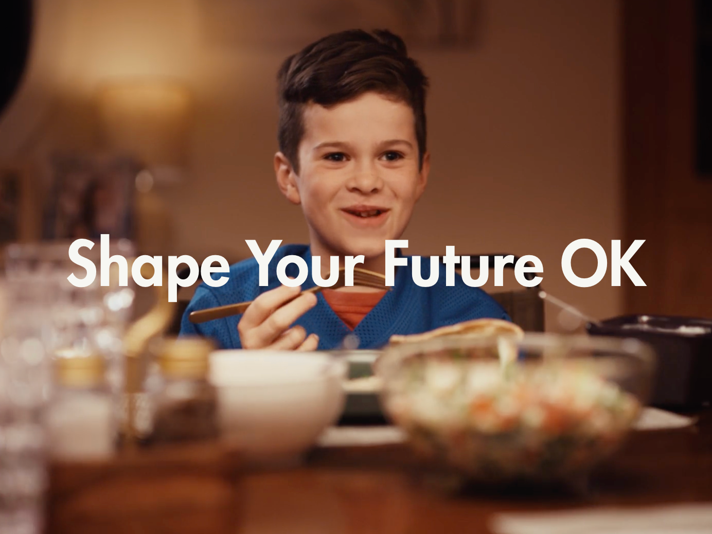A young boy with short dark hair smiling while eating with chopsticks at a table with various dishes, with a caption that reads 'Shape Your Future OK.'