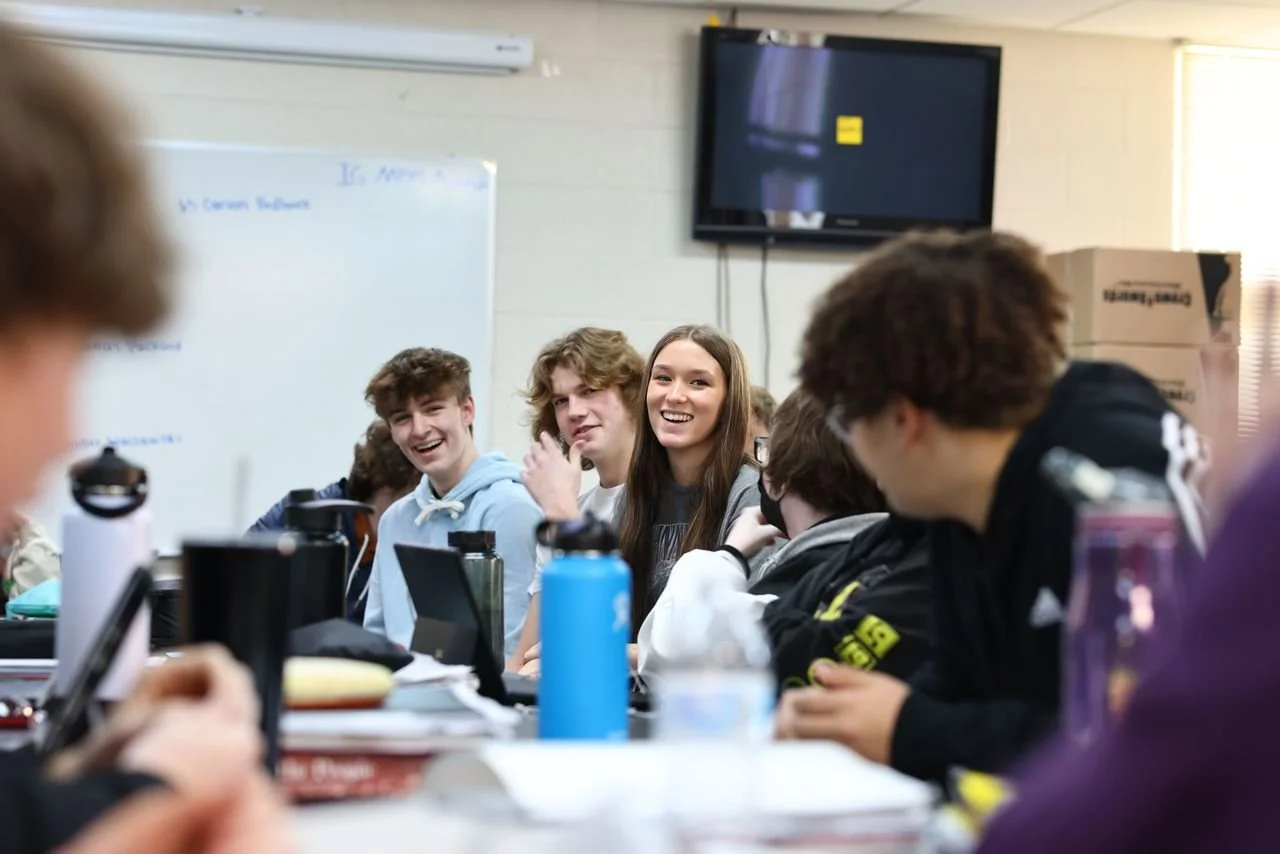 A classroom scene with students sitting at a table, smiling and engaging with each other, with a whiteboard and a TV on the wall in the background.