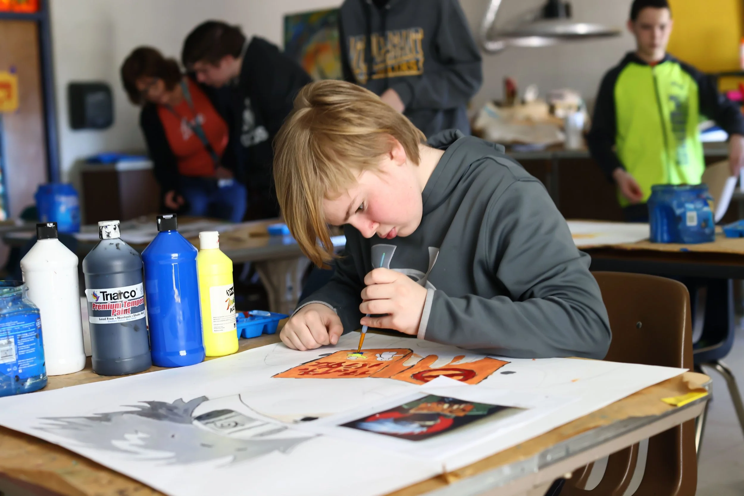 A young boy with red hair and a gray hoodie displays concentration as he works on a colorful paper art project at a table with bottles of paint and brushes, in a classroom setting with other children and adults in the background.