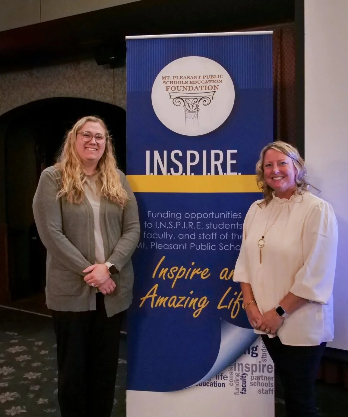 Two women standing next to a Mt. Pleasant Public Schools Foundation banner at an indoor event. They are smiling and dressed in business casual attire.