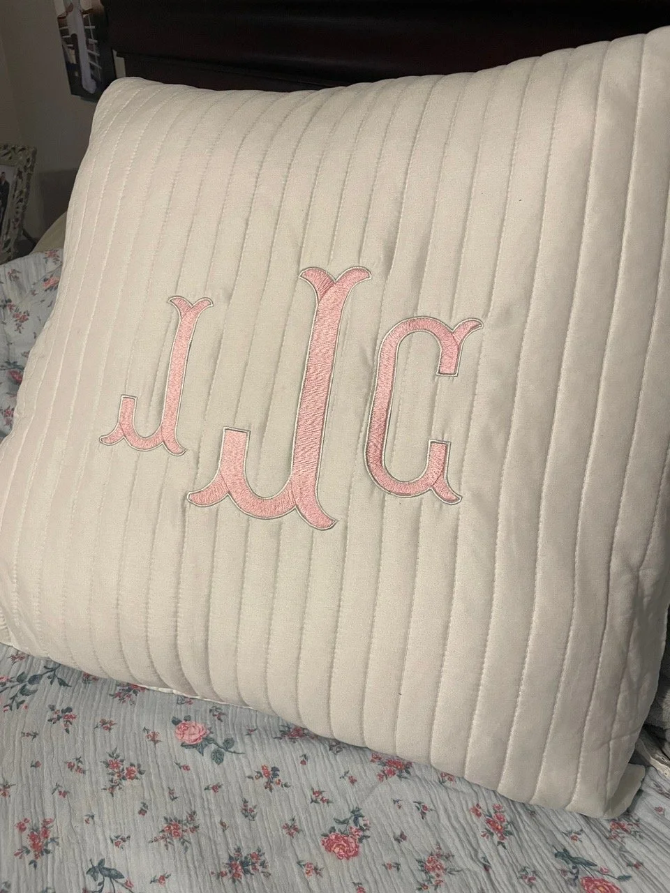 A white cushion with vertical quilted lines and pink embroidered initials 'JLC' resting on a floral-patterned bedspread.