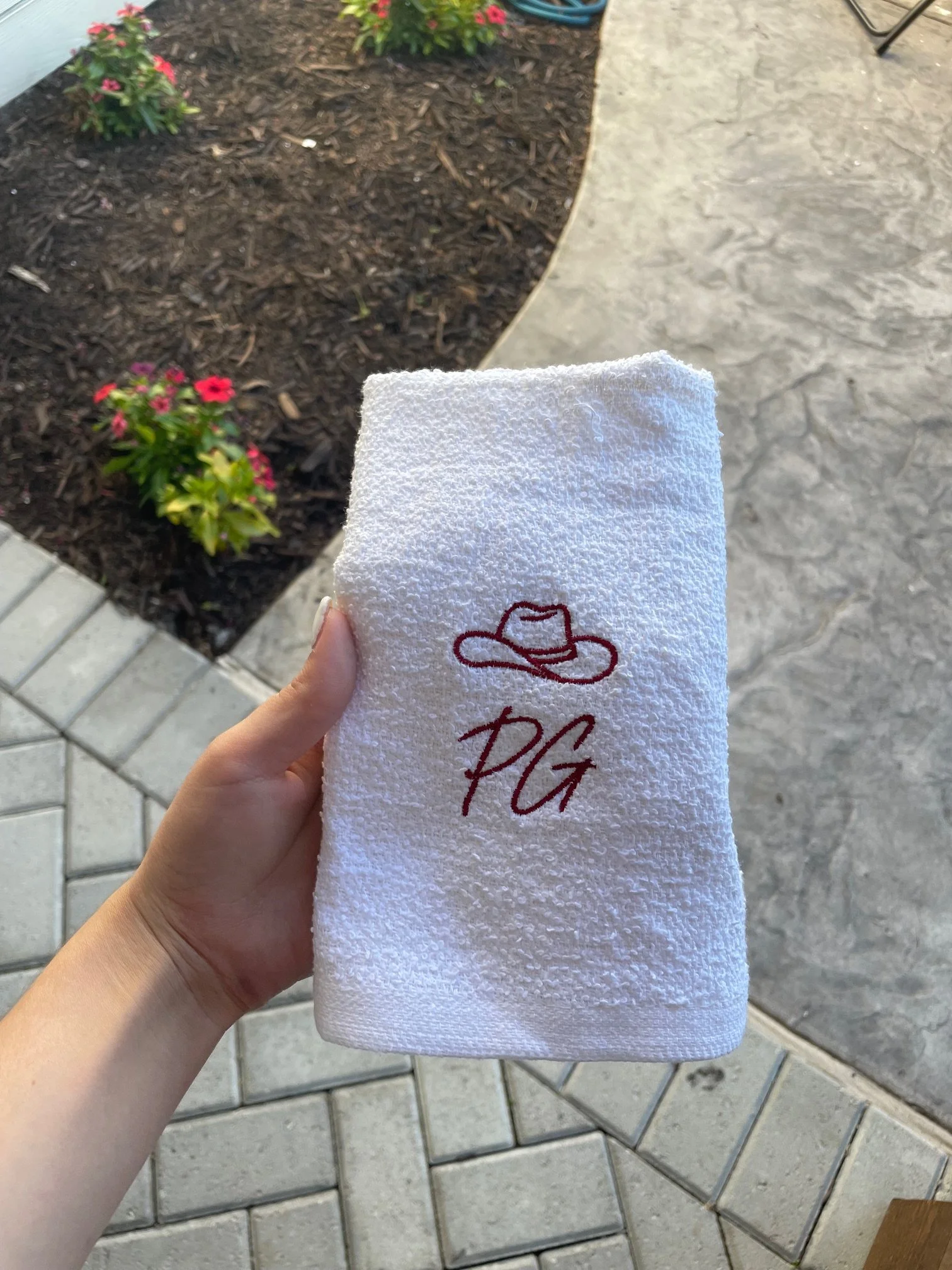 A hand holding a white towel with red embroidered writing and a hat design, outside on a stone patio next to a flower bed with pink and red flowers and mulch.