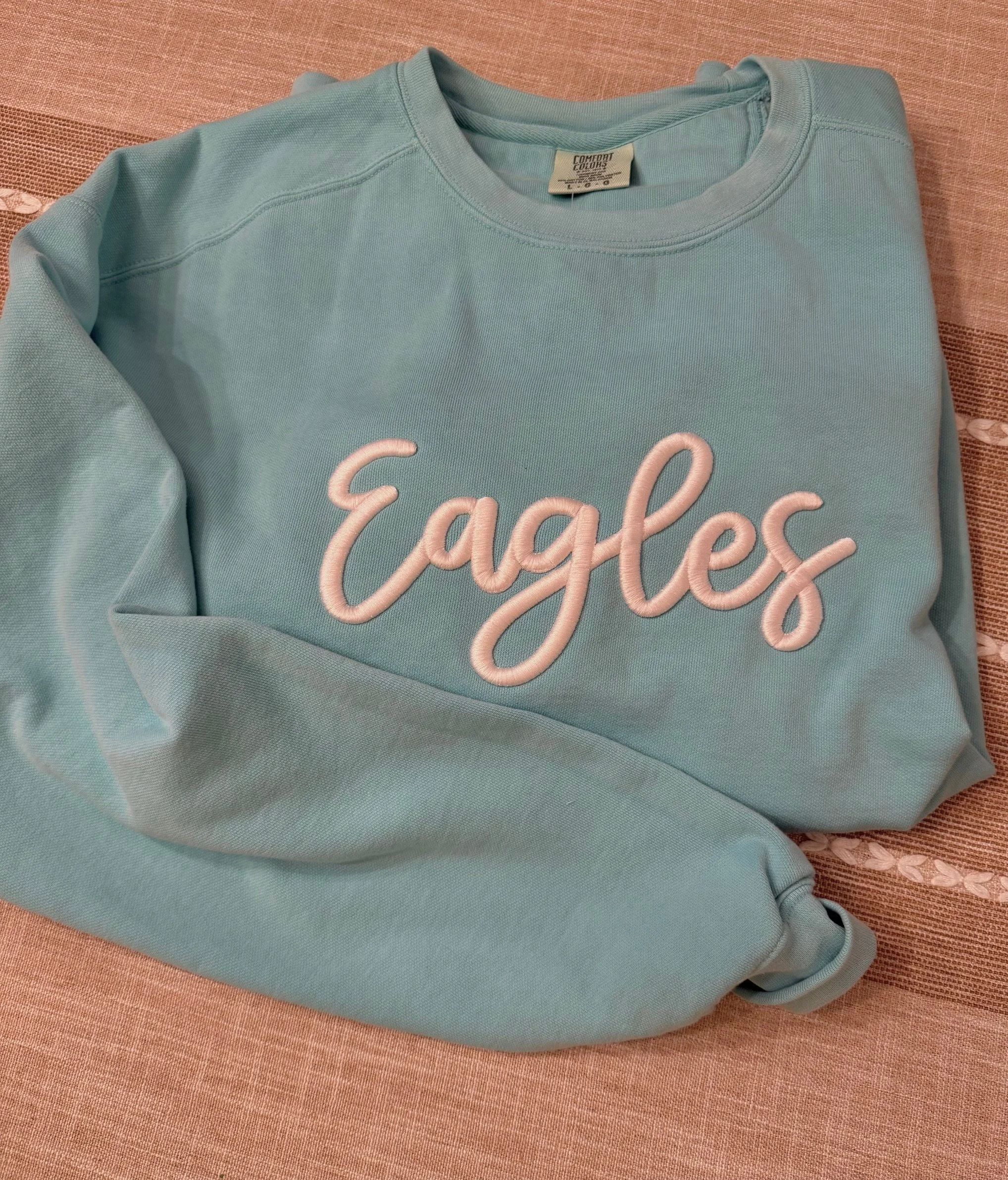 Light blue sweatshirt with the word 'Cackle' embroidered in pink on the front, resting on a pink fabric surface.