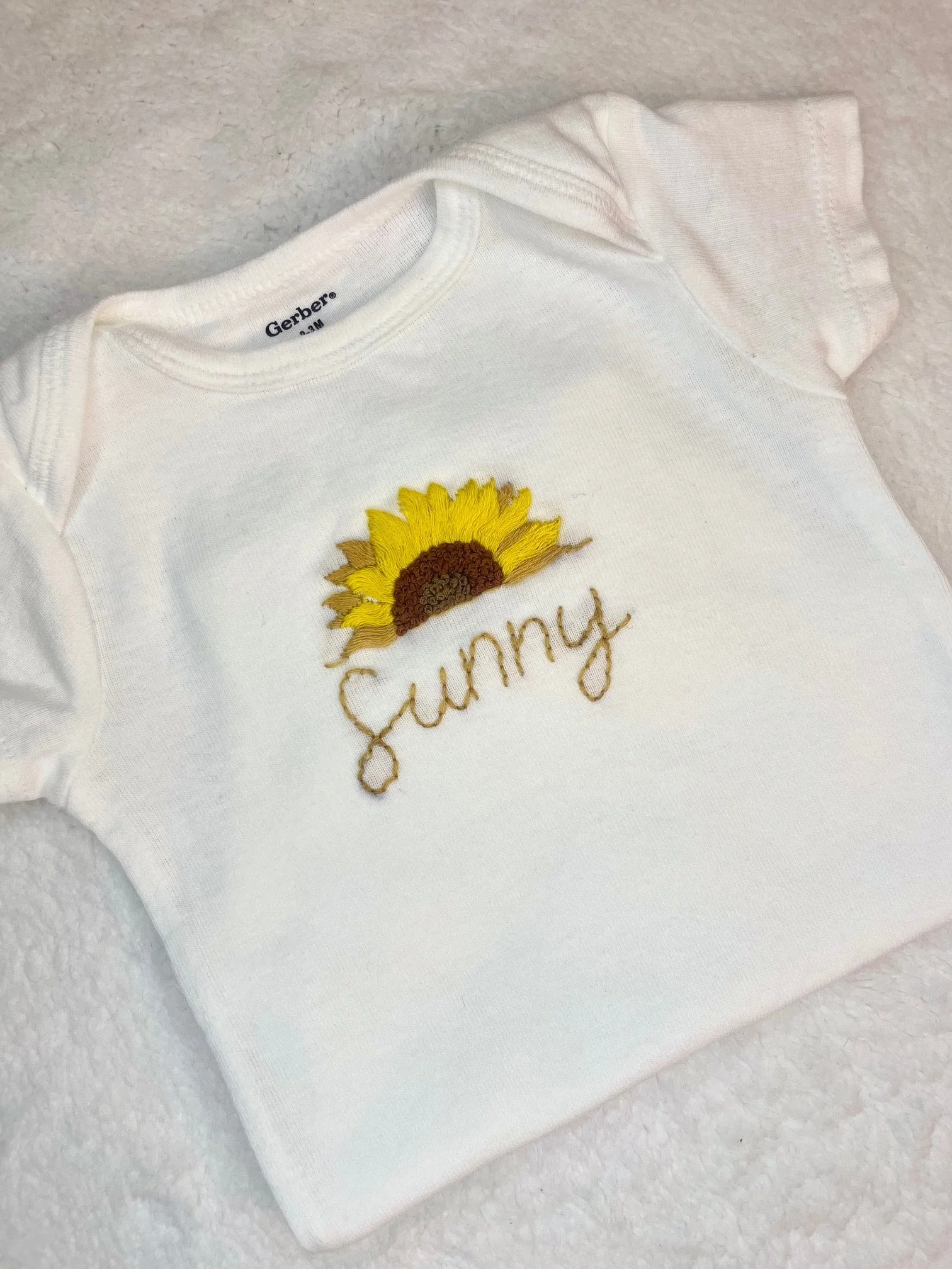 White baby onesie with embroidered sunflower and the words "sunny" on a soft, textured surface.