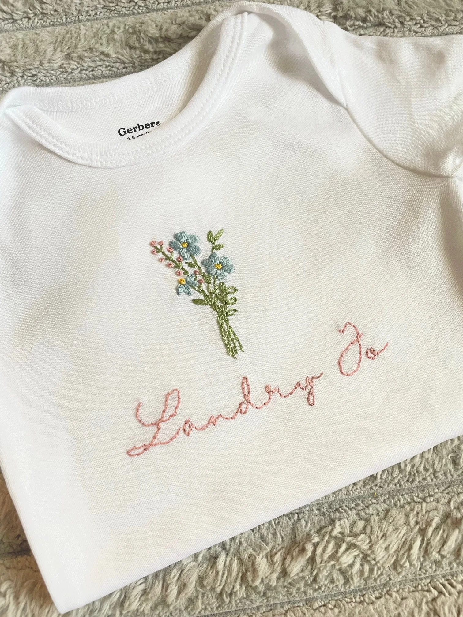 White Gerber t-shirt with embroidered floral design and pink cursive text, lying on a gray textured blanket.