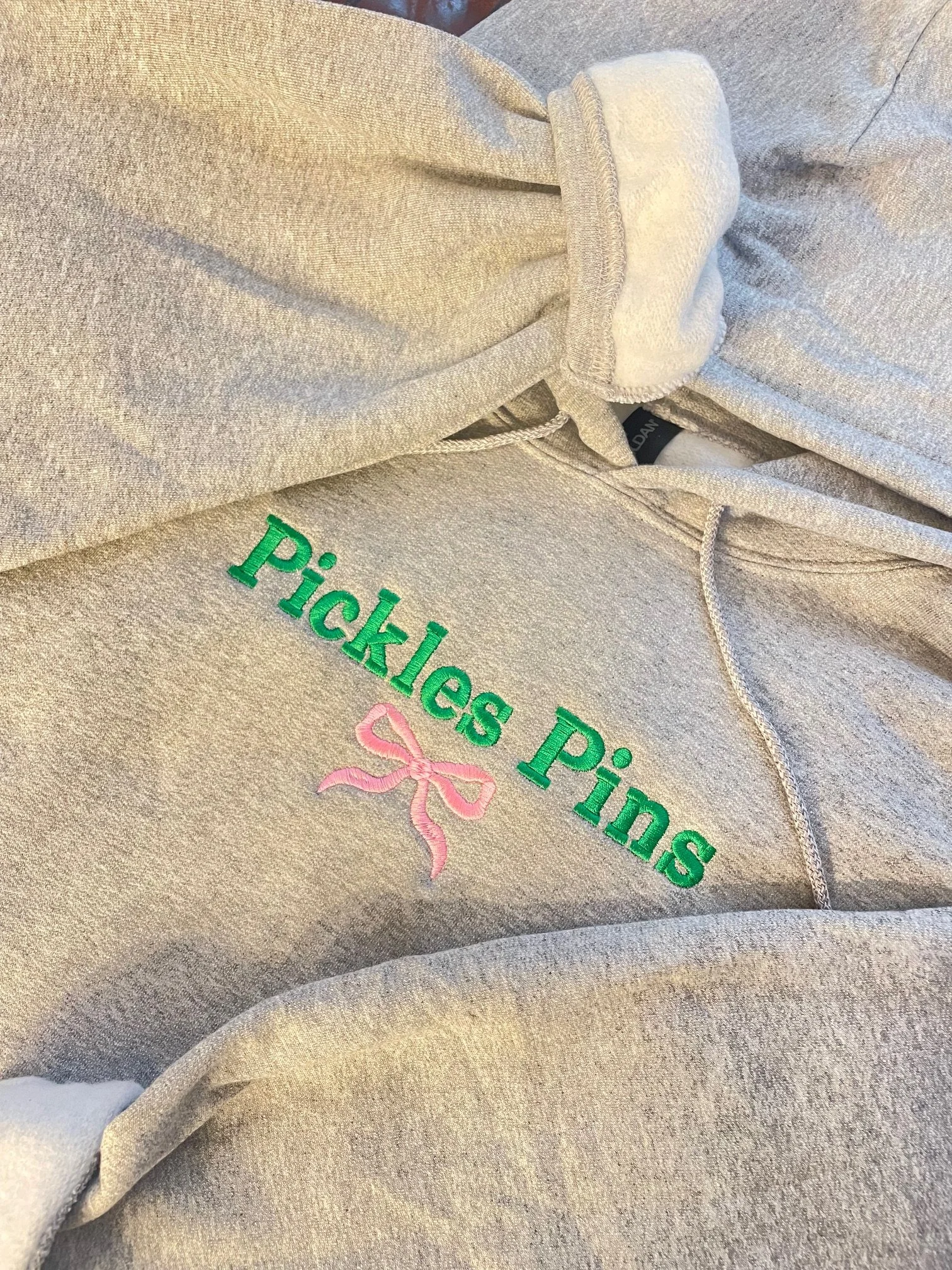 Close-up of a beige hoodie with green embroidered text that says 'Pickles Pinks' and a pink embroidered bow.