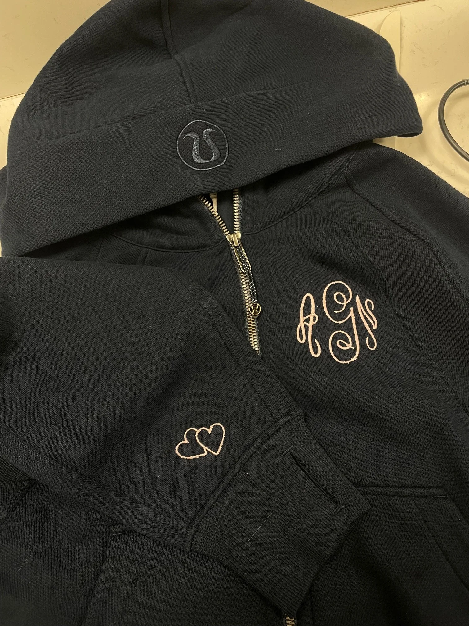 A black hoodie with embroidered gold hearts on the sleeve and an embroidered monogram on the chest, partially zipped open, lying on a beige surface.