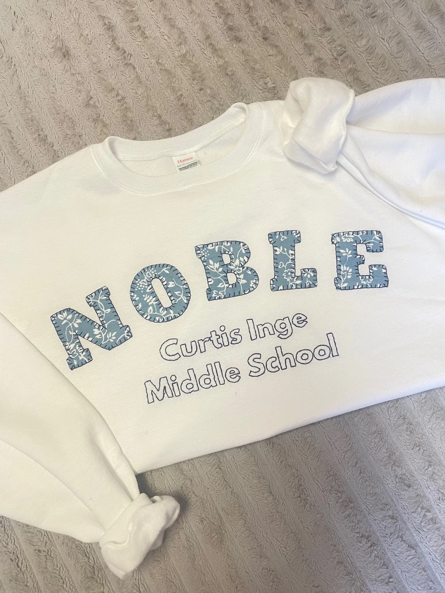 White t-shirt with blue floral reading 'MIDDLE' and 'Curtis Middle School' placed on a textured surface with a white rolled-up sleeve partially visible.